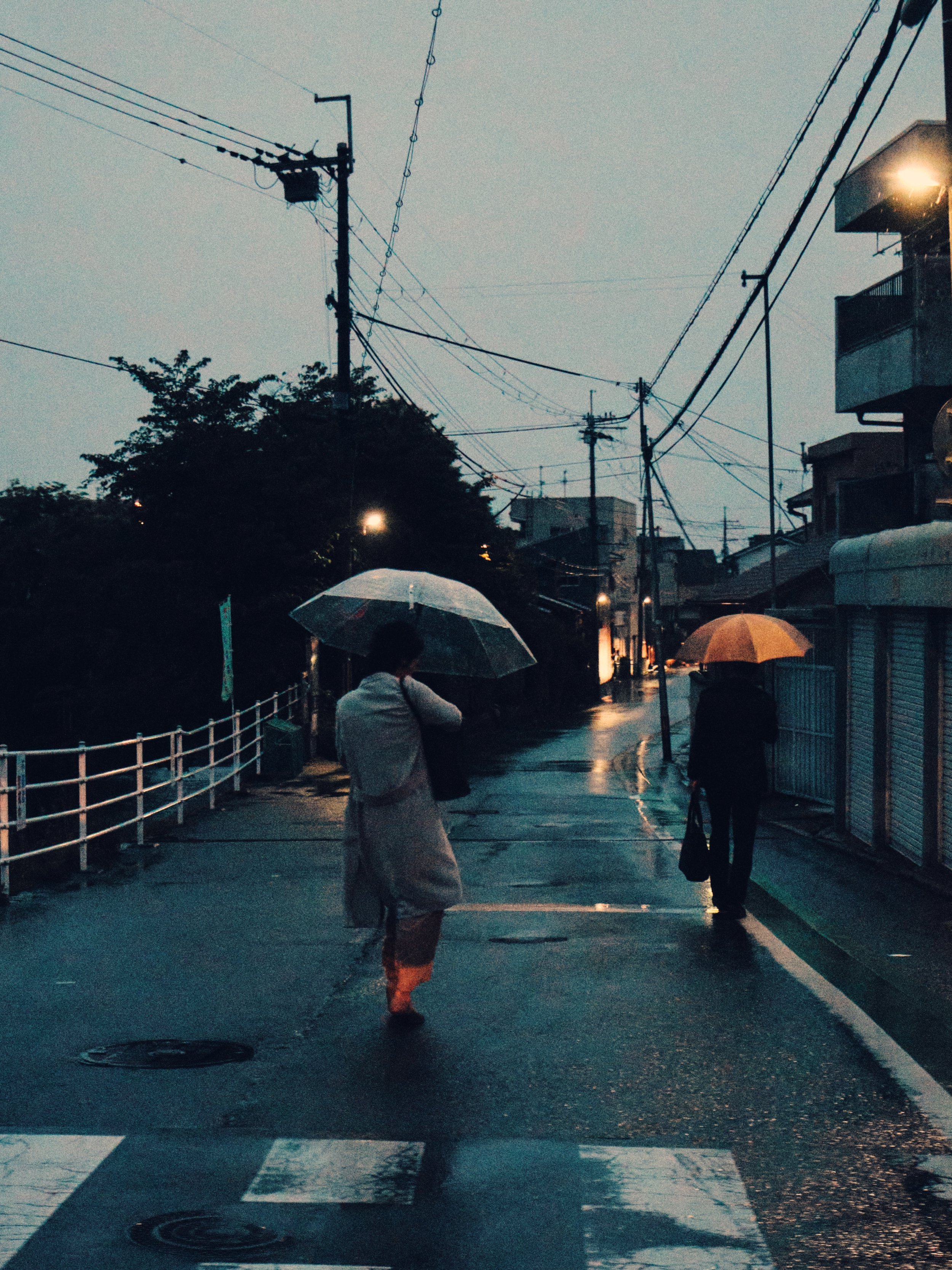 A rainy evening in a quiet urban street with two people walking with umbrellas. One person, in a light coat, stands near a crosswalk, while the other walks away, carrying a bag. The street is wet and illuminated by streetlights, with buildings and po