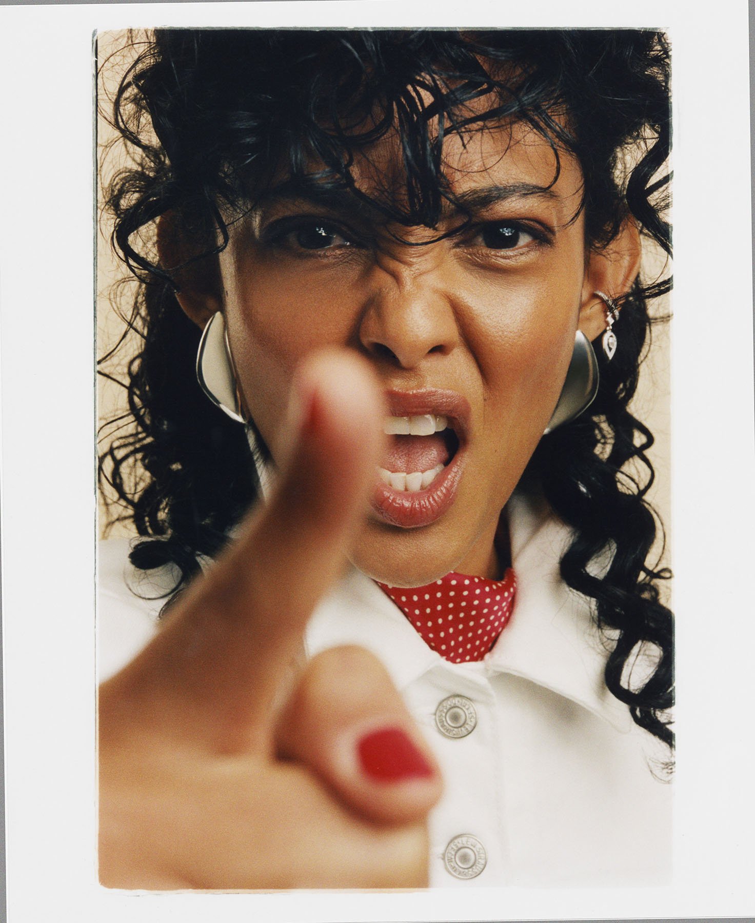 Woman with curly black hair, wearing earrings and a white shirt with red polka dot necktie, making an angry face and pointing directly at the camera.