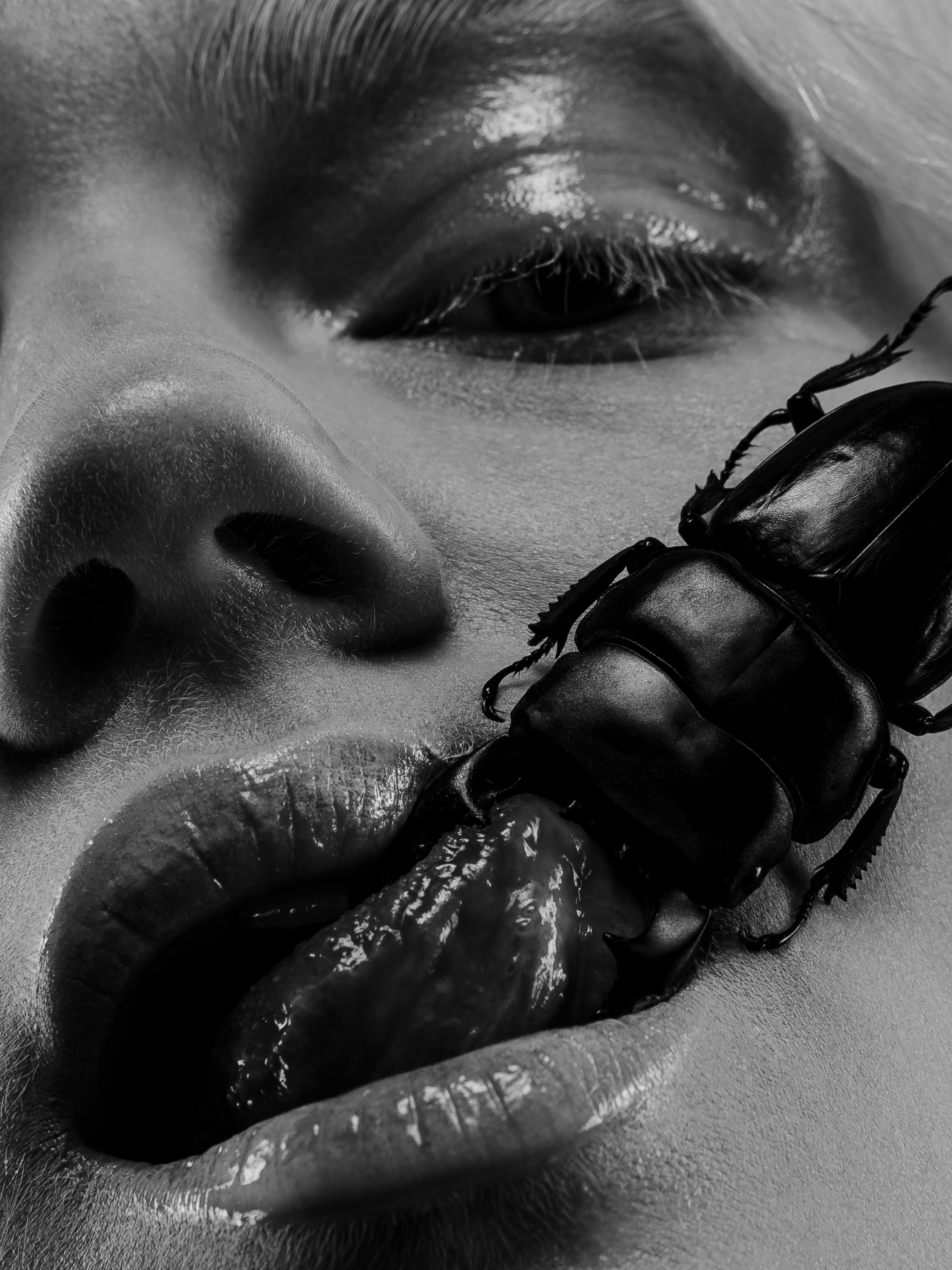 A close-up black and white photo of a person's face showing a nose, part of an eye, and lips, with a large beetle on the tongue.