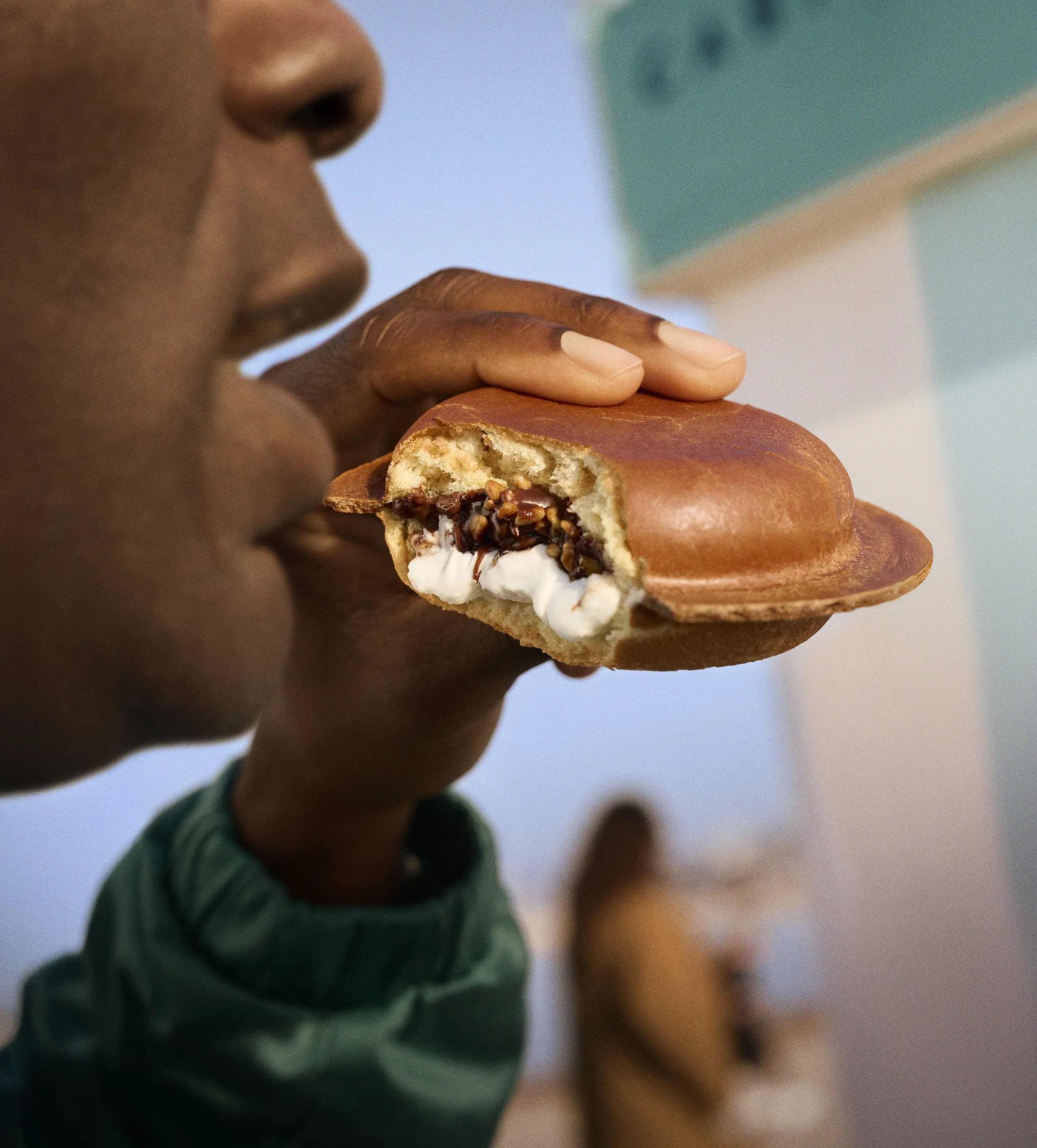 Person holding and taking a bite out of a sandwich-like food item with a filling of whipped cream and chopped nuts or chocolate.