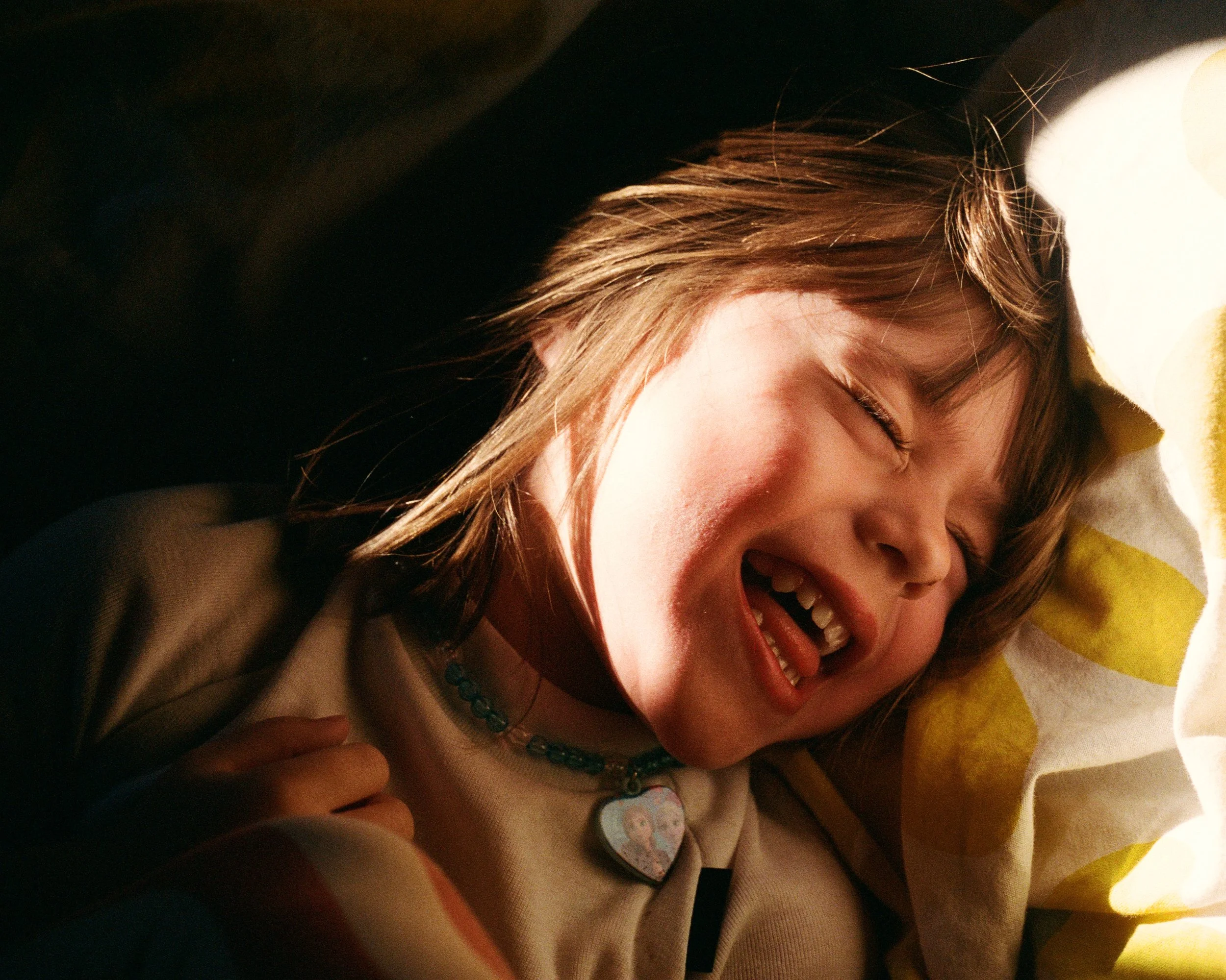 A young girl with brown hair is lying on her side, smiling and laughing with her eyes closed, resting her head on a yellow, patterned pillow.
