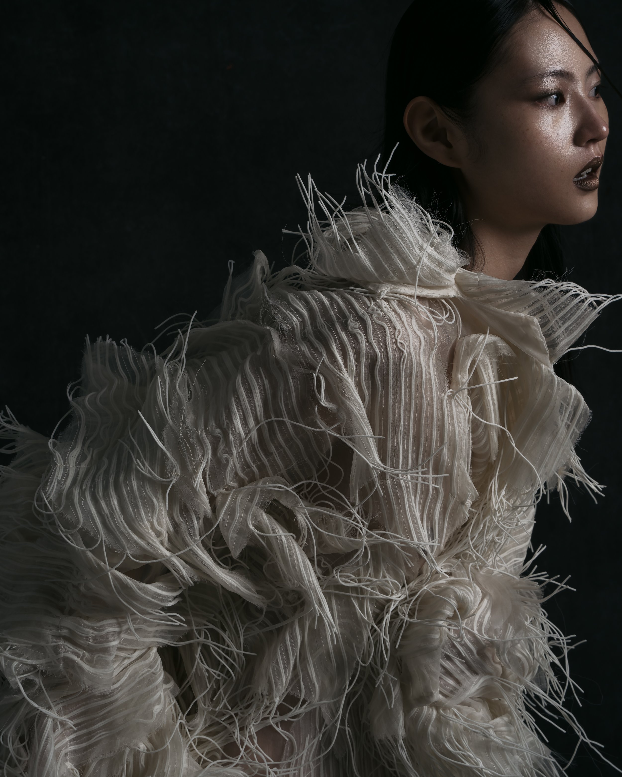A woman with dark hair and makeup wearing an avant-garde cream-colored garment made of layered, textured fabric with hanging threads, set against a dark background.