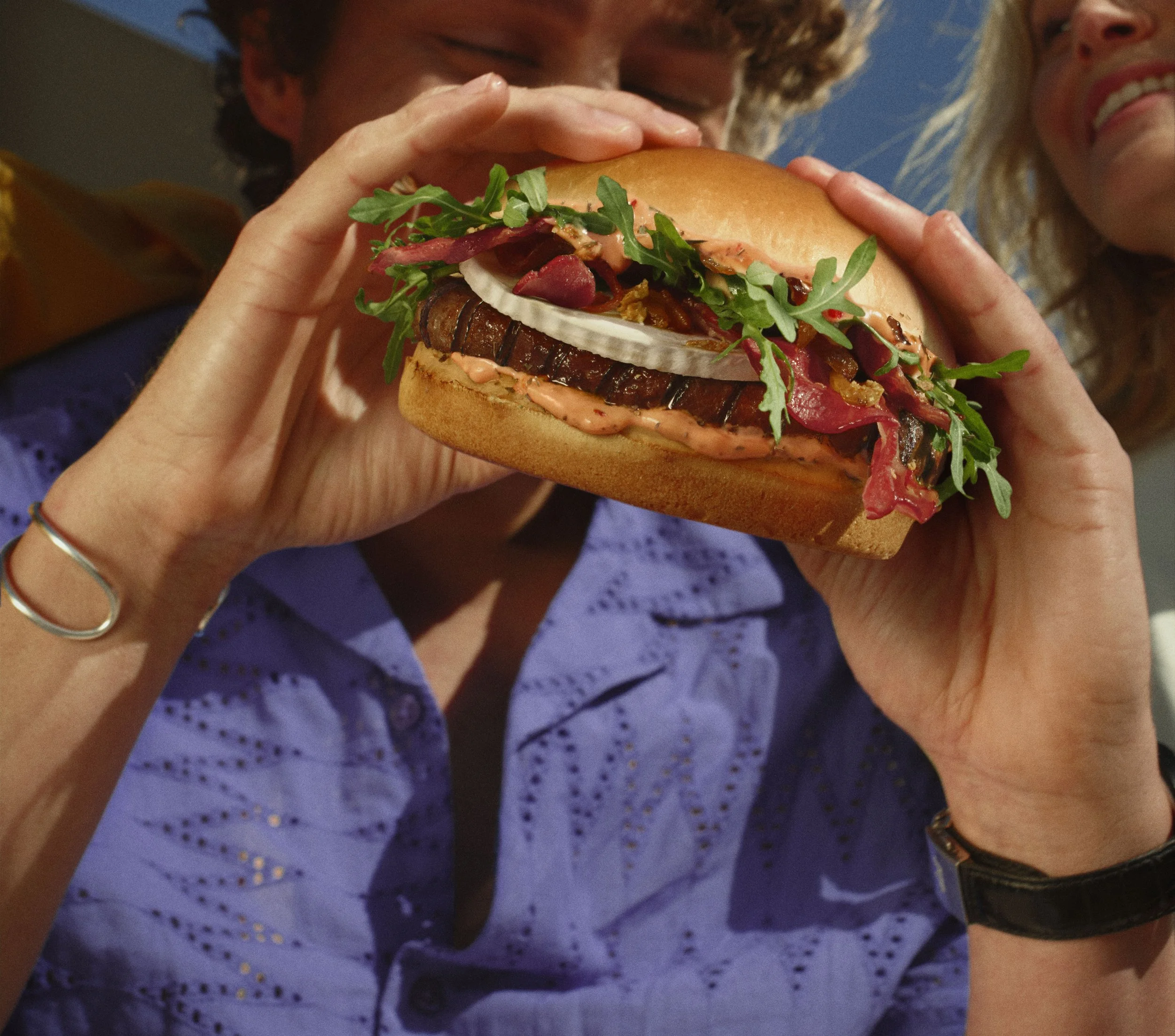 A person holding a sandwich with grilled eggplant, fresh greens, tomato, onion, and sauce, with another person smiling in the background.