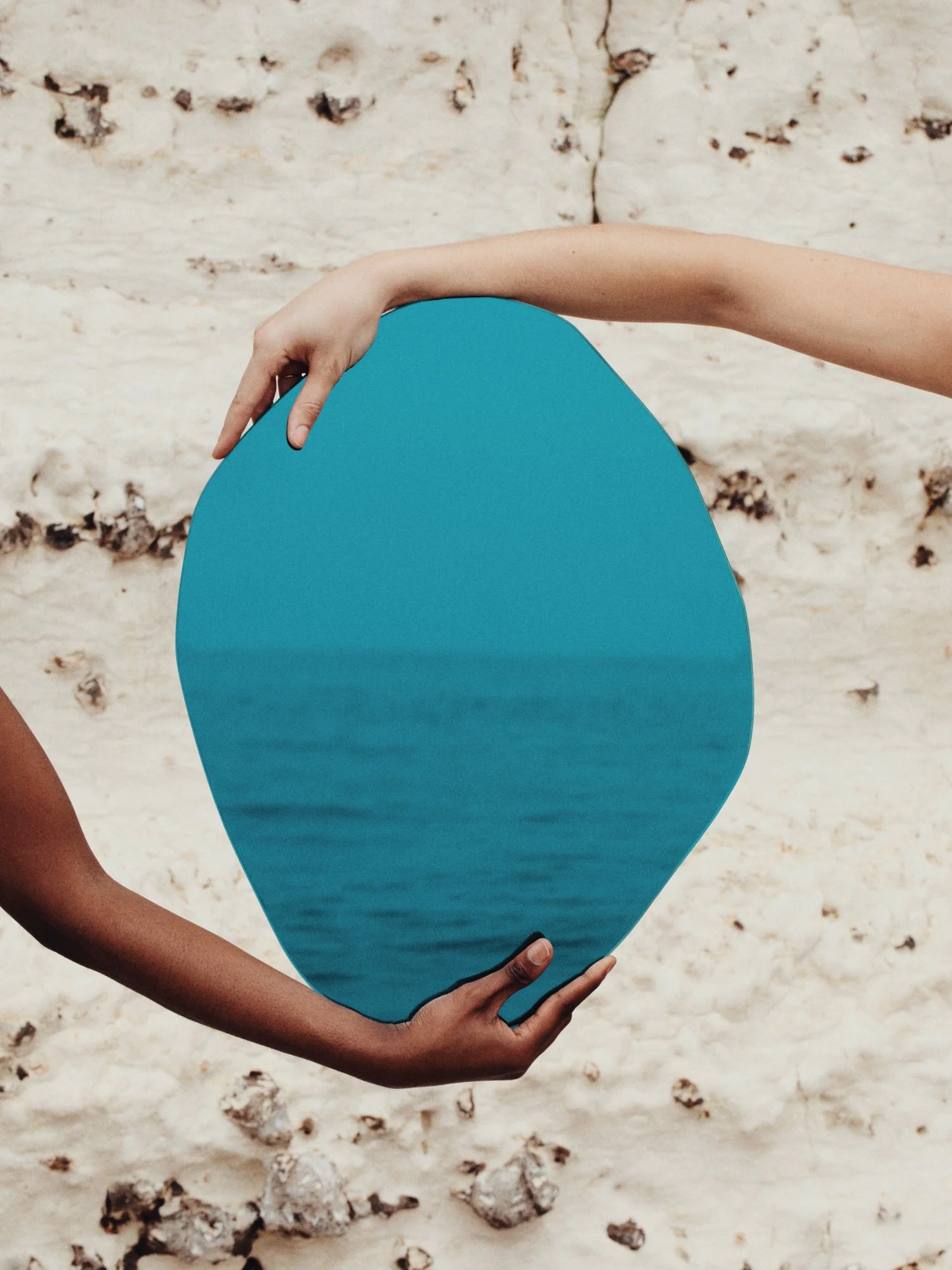 Two hands holding a blue, oval-shaped object with a water or ocean scene reflected on its surface, against a sandy background.