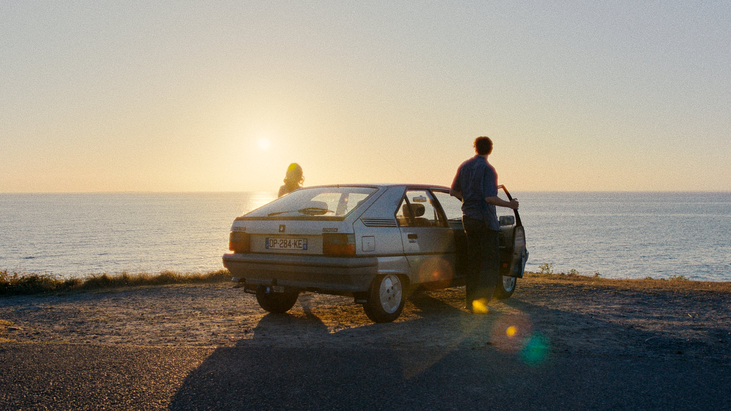 A man standing outside a parked car by the ocean during sunset, with a woman visible through the car window.