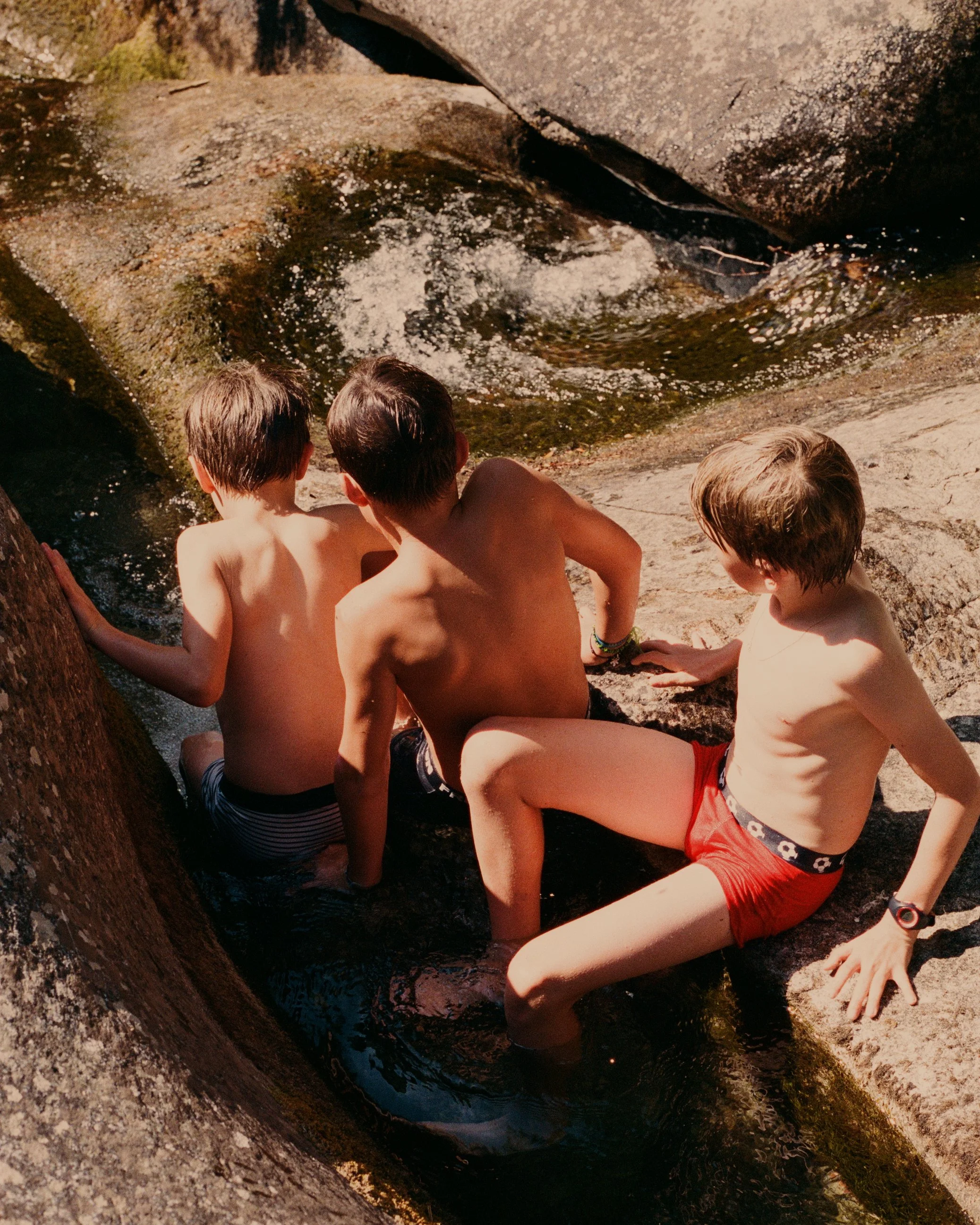 Three boys sitting on rocks near a small waterfall or stream, in a natural outdoor setting.
