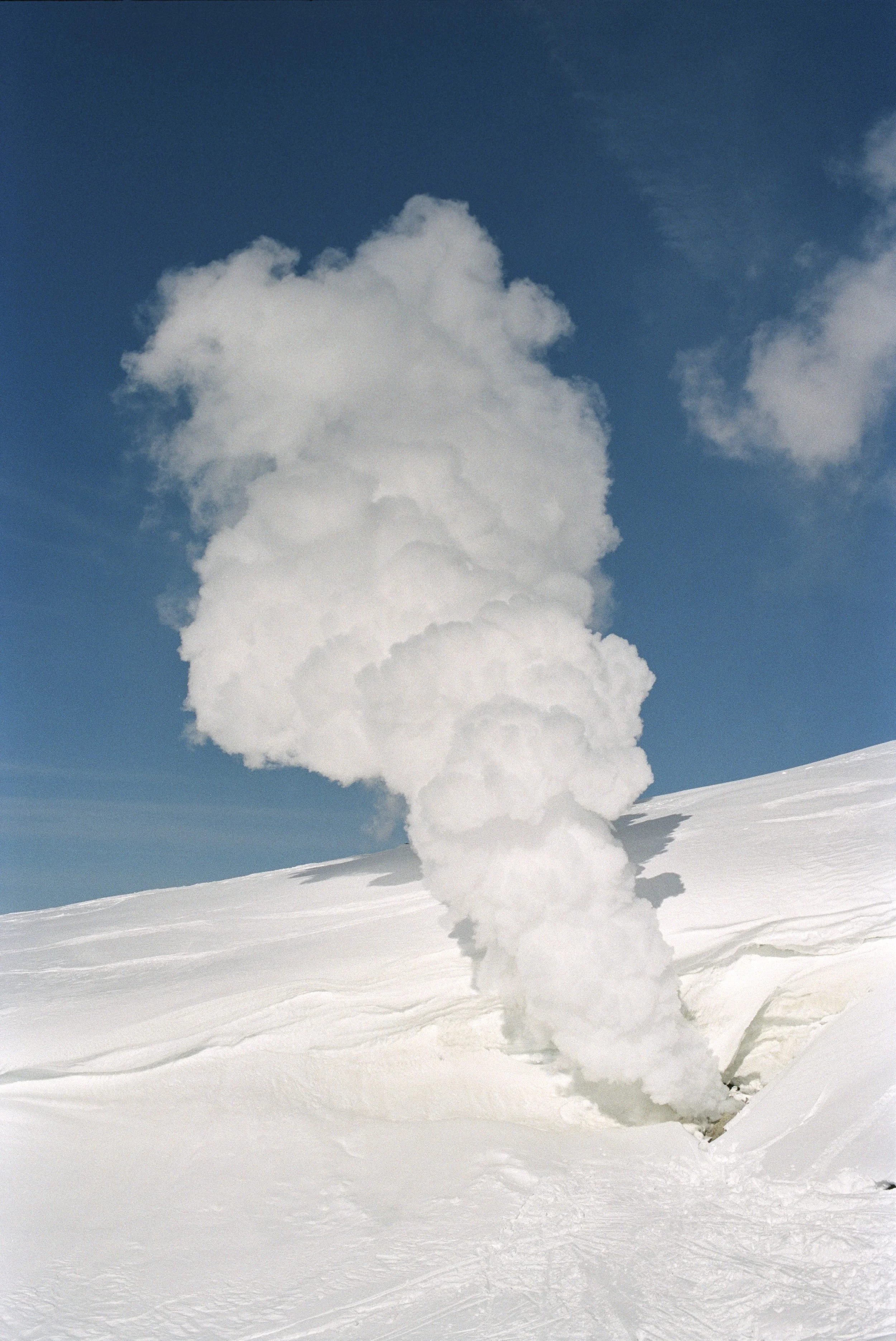 A plume of white smoke or steam rising from a snowy landscape against a blue sky with a few clouds.