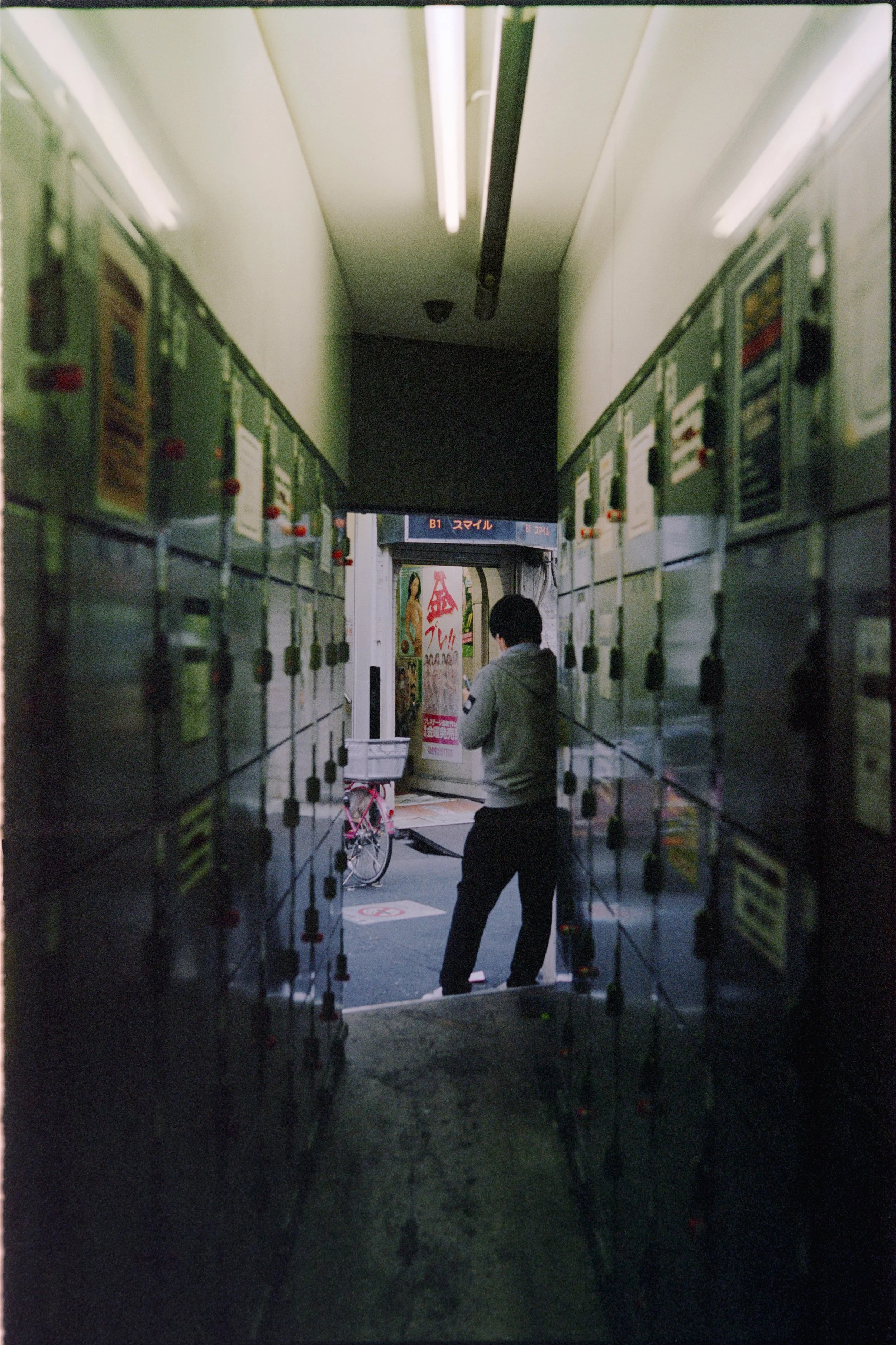 A narrow hallway with green lockers on both sides and fluorescent lighting on the ceiling, leading to an outdoor entrance where a person is standing and looking at their phone.