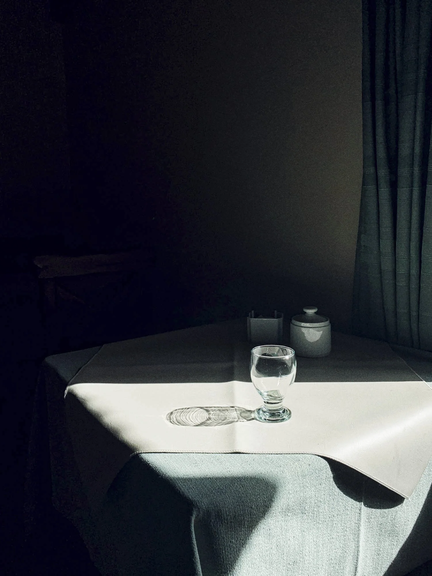 Sunlight cast shadows on a table with a glass filled with water, a white napkin, and small containers, in a dimly lit room with dark walls and curtains.
