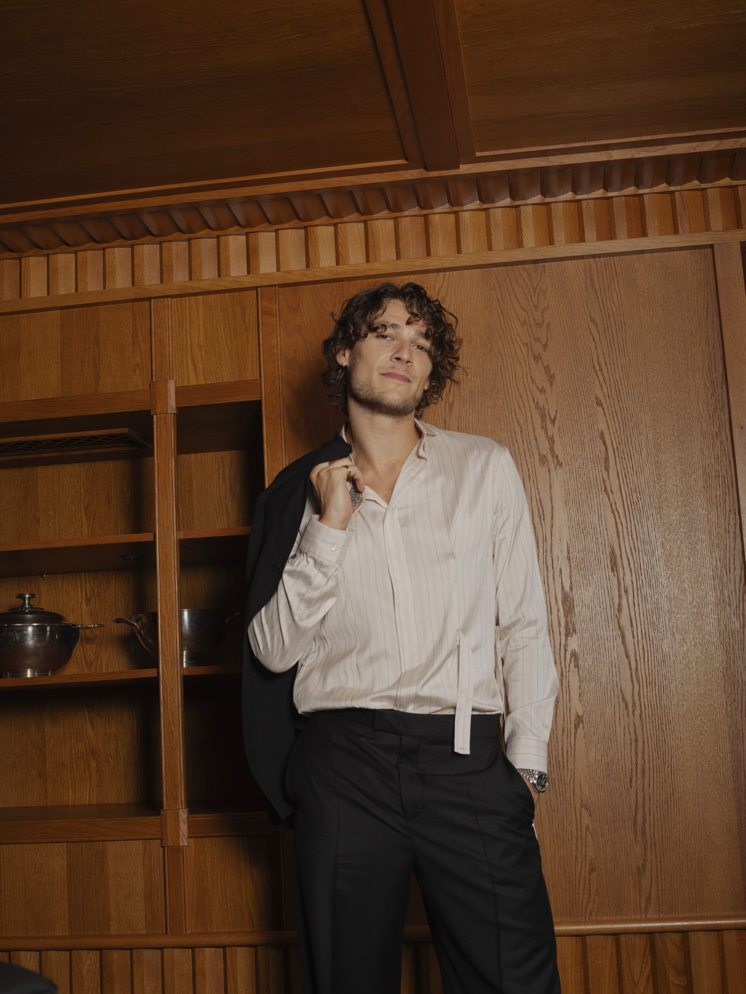 A young man with curly hair in a white shirt and black pants, carrying a black blazer over his shoulder, leaning against a wooden wall in a room with wood-paneled walls and shelves with metal dishes.