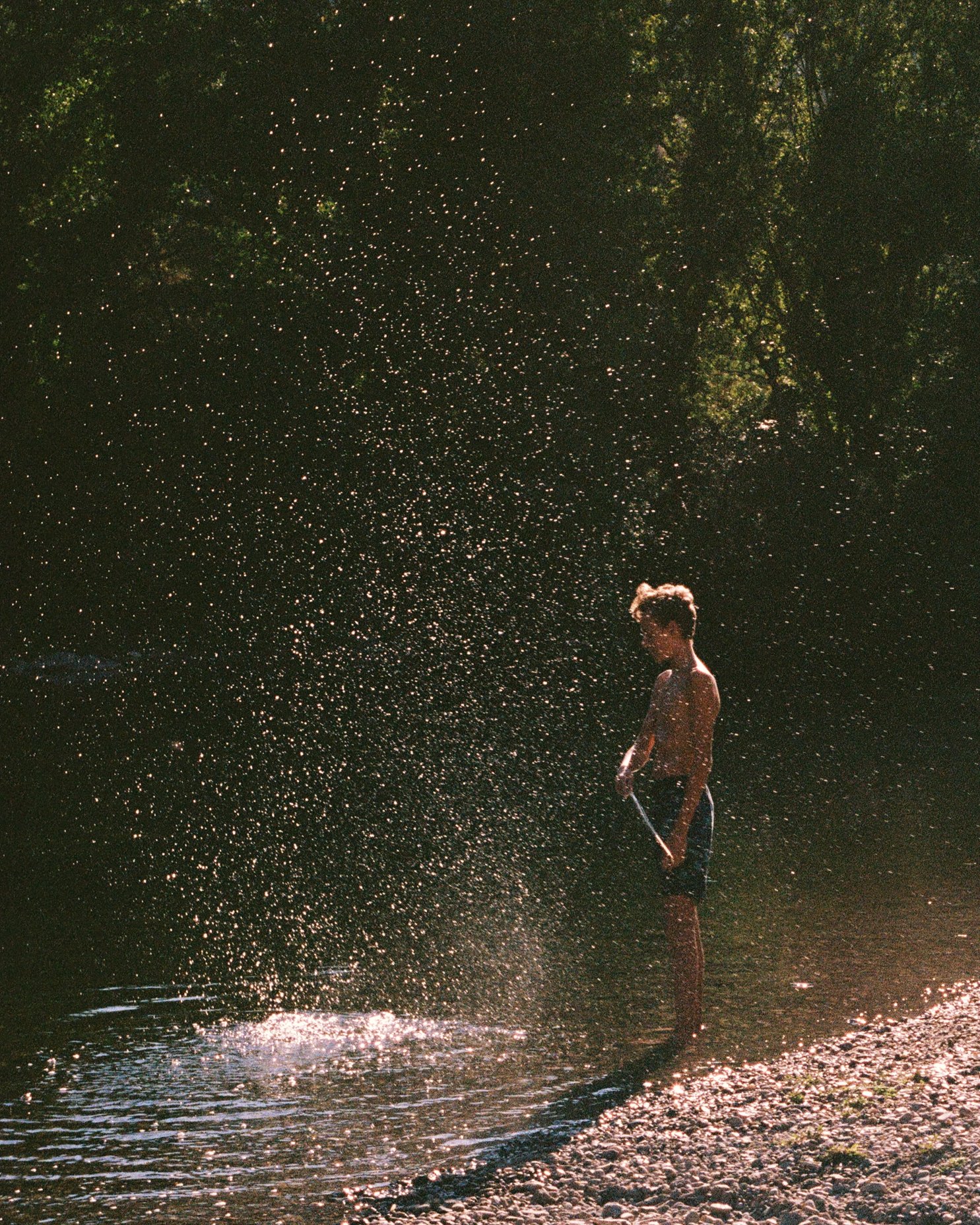 A young person throwing water into the air at a riverbank during sunset or sunrise, with sunlight causing the water droplets to sparkle, and trees in the background.