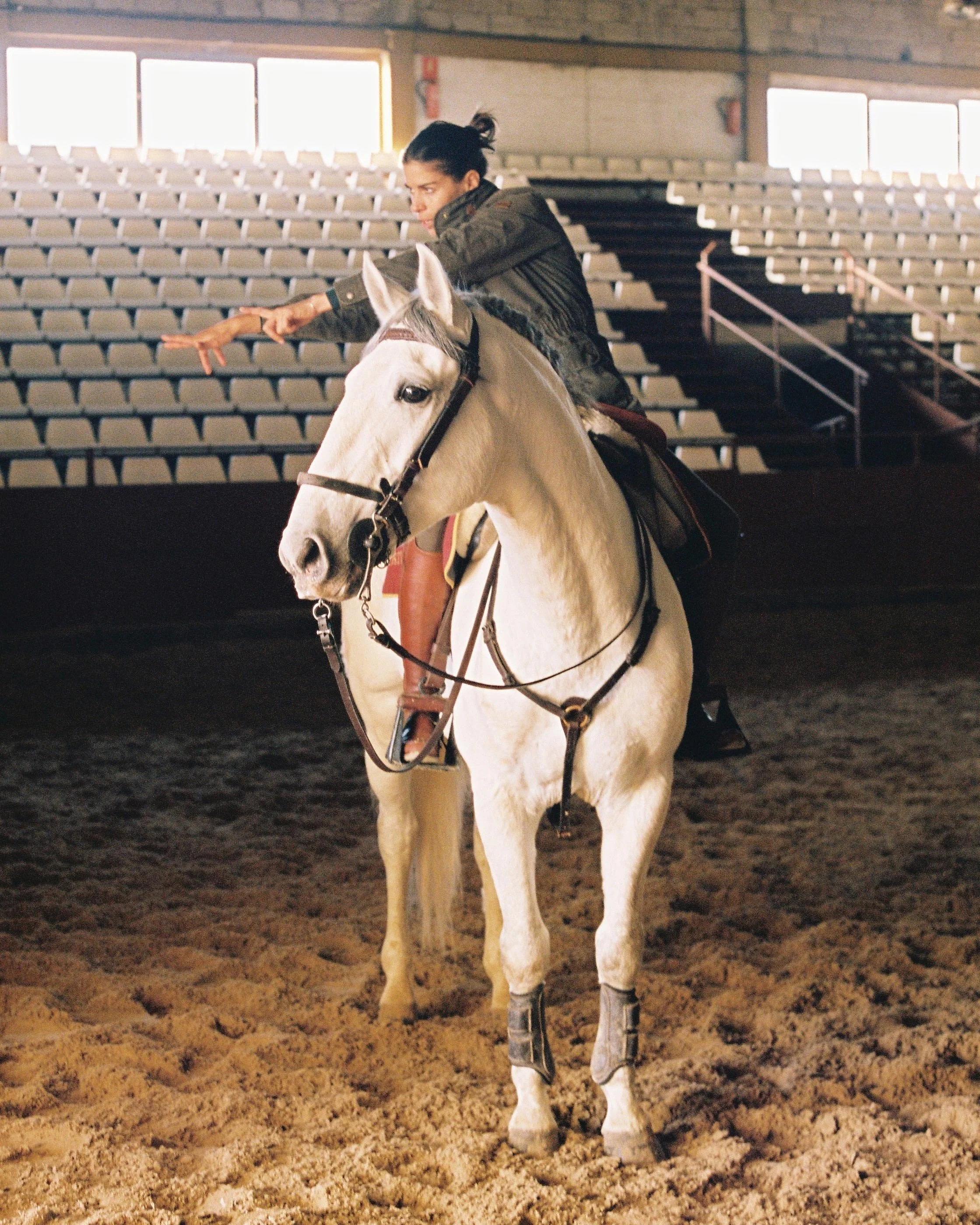 A woman riding a white horse inside a rodeo arena with sandy ground, empty bleachers, and windows in the background.