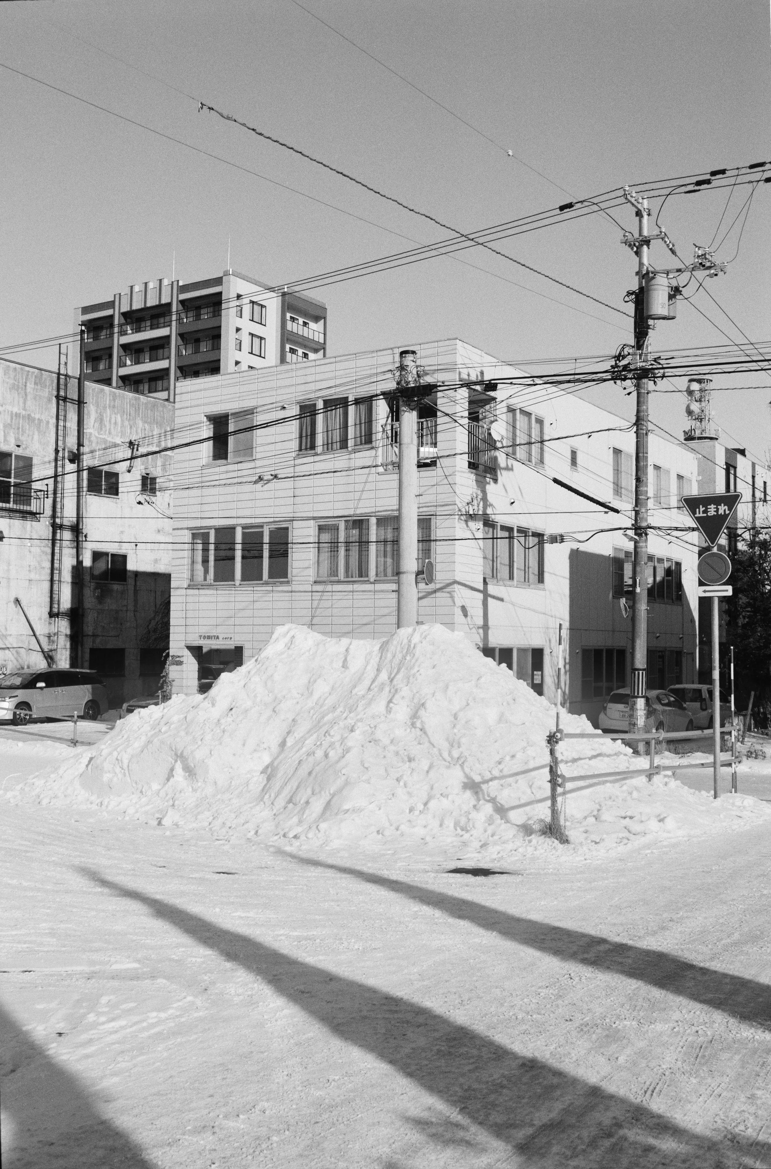 Black and white photo of a snow mound next to a utility pole, with buildings, cars, and power lines in the background.