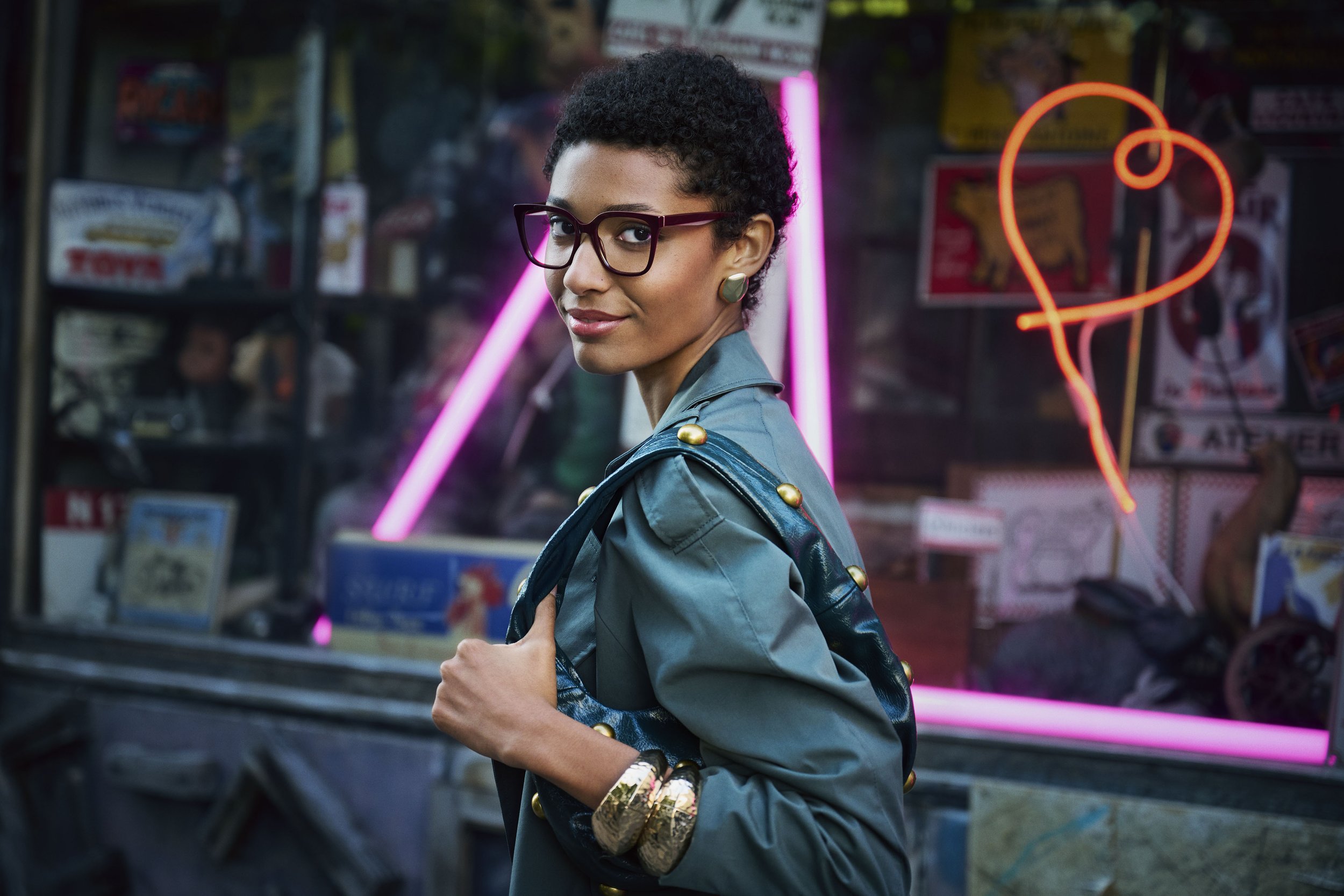A woman with short, curly black hair and glasses stands in front of a shop window with neon lights, holding a bag. She wears a teal jacket with gold studs on the shoulders and has gold bracelets on her wrist, with pink neon and a heart-shaped neon si
