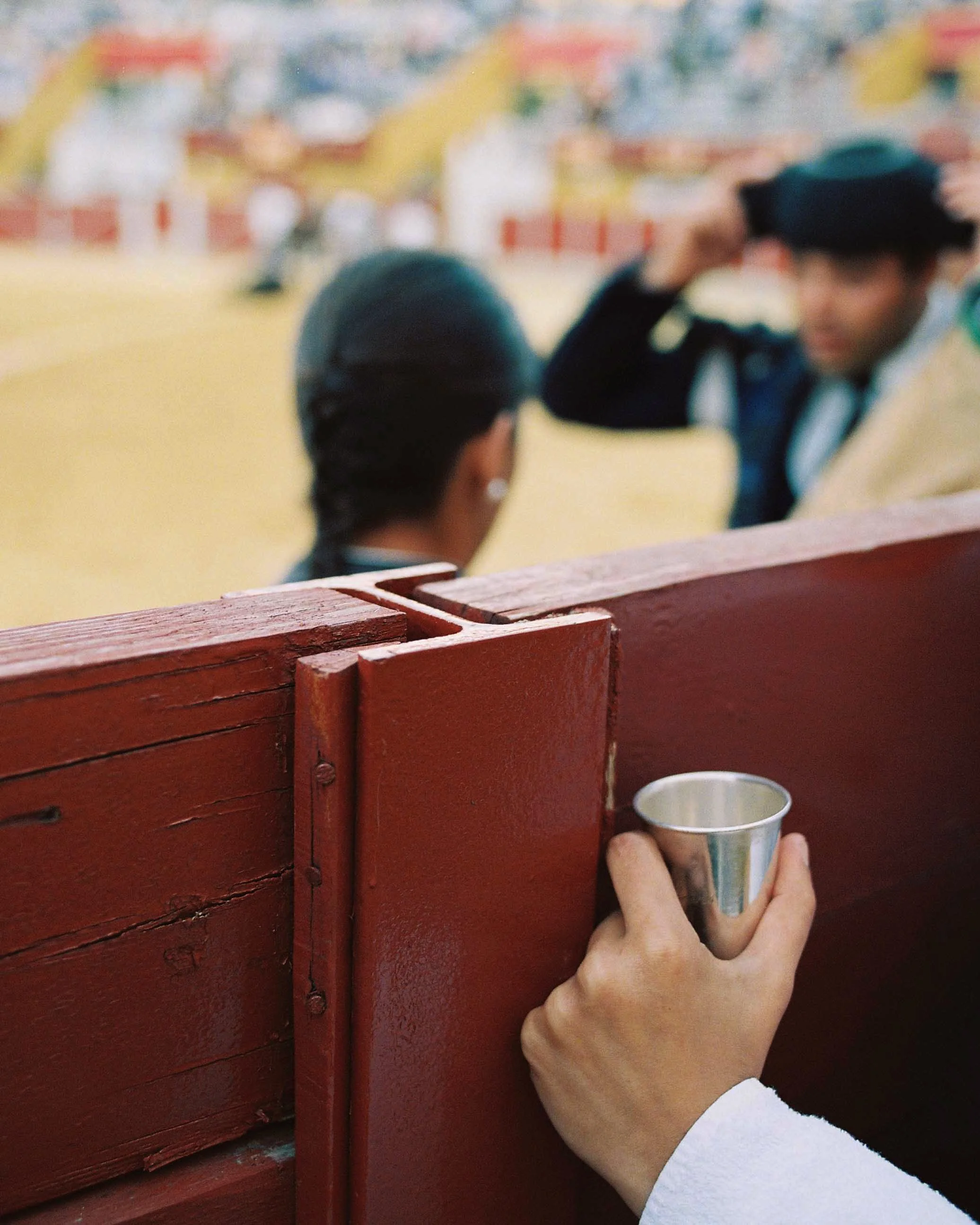 Close-up of a hand holding a small metal cup peeking over a red wooden barrier, with two people in traditional attire and a performer in a hat in the background at an outdoor event.