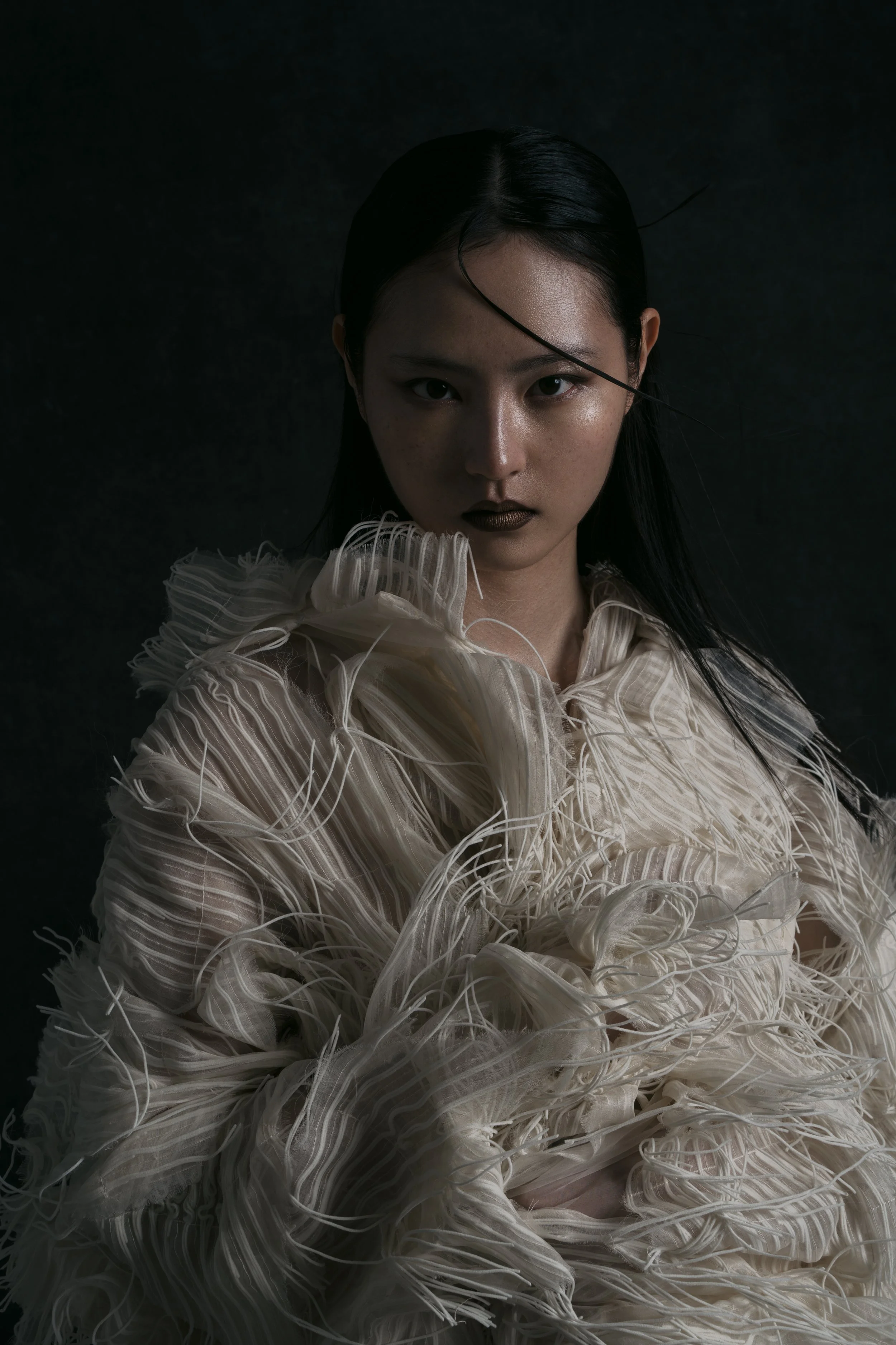 A woman with dark hair, wearing dark lipstick, and a textured cream-colored garment with feather-like details, posed against a dark background.