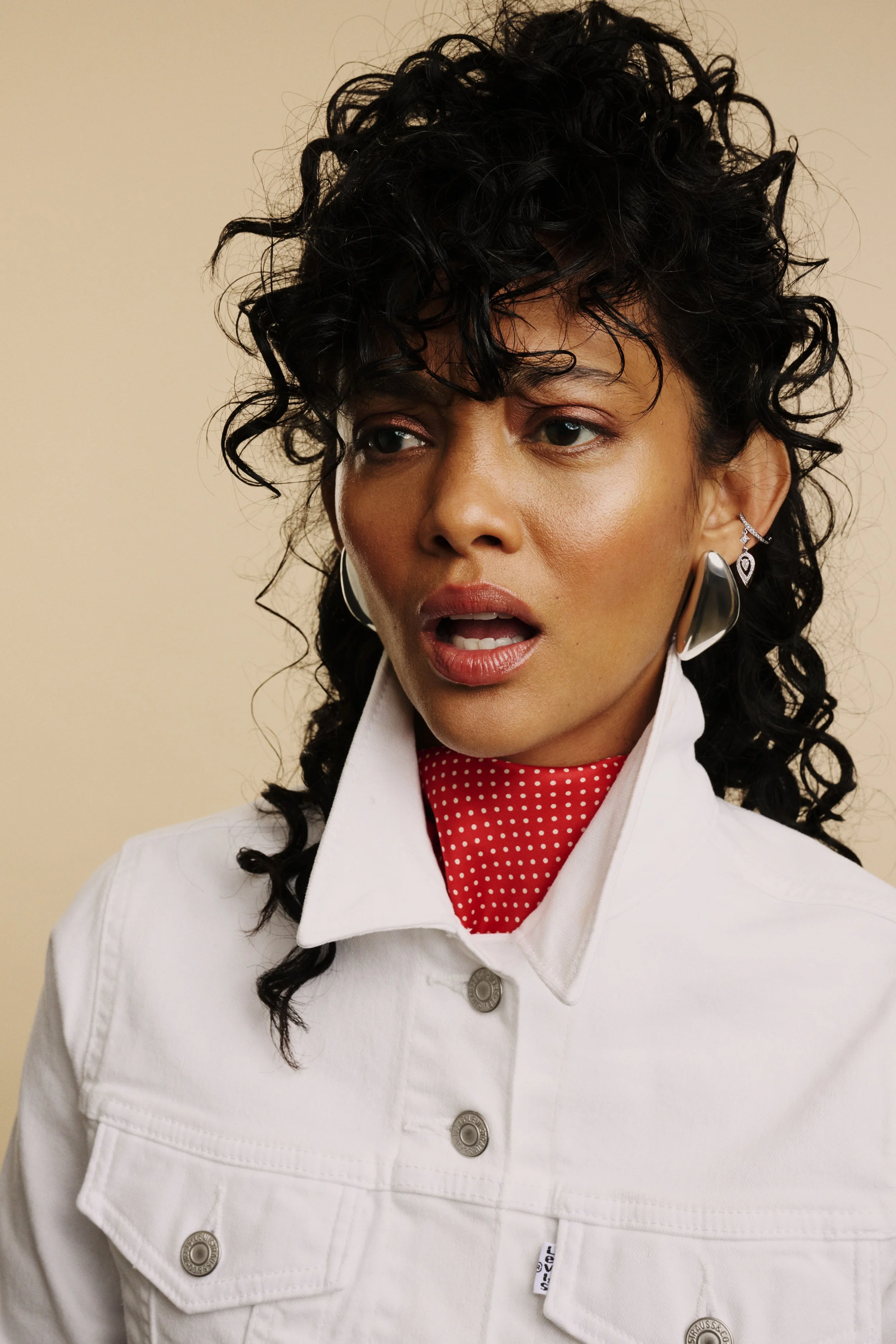 A woman with curly black hair and earrings, wearing a white denim jacket over a red polka dot shirt, appears surprised or confused.