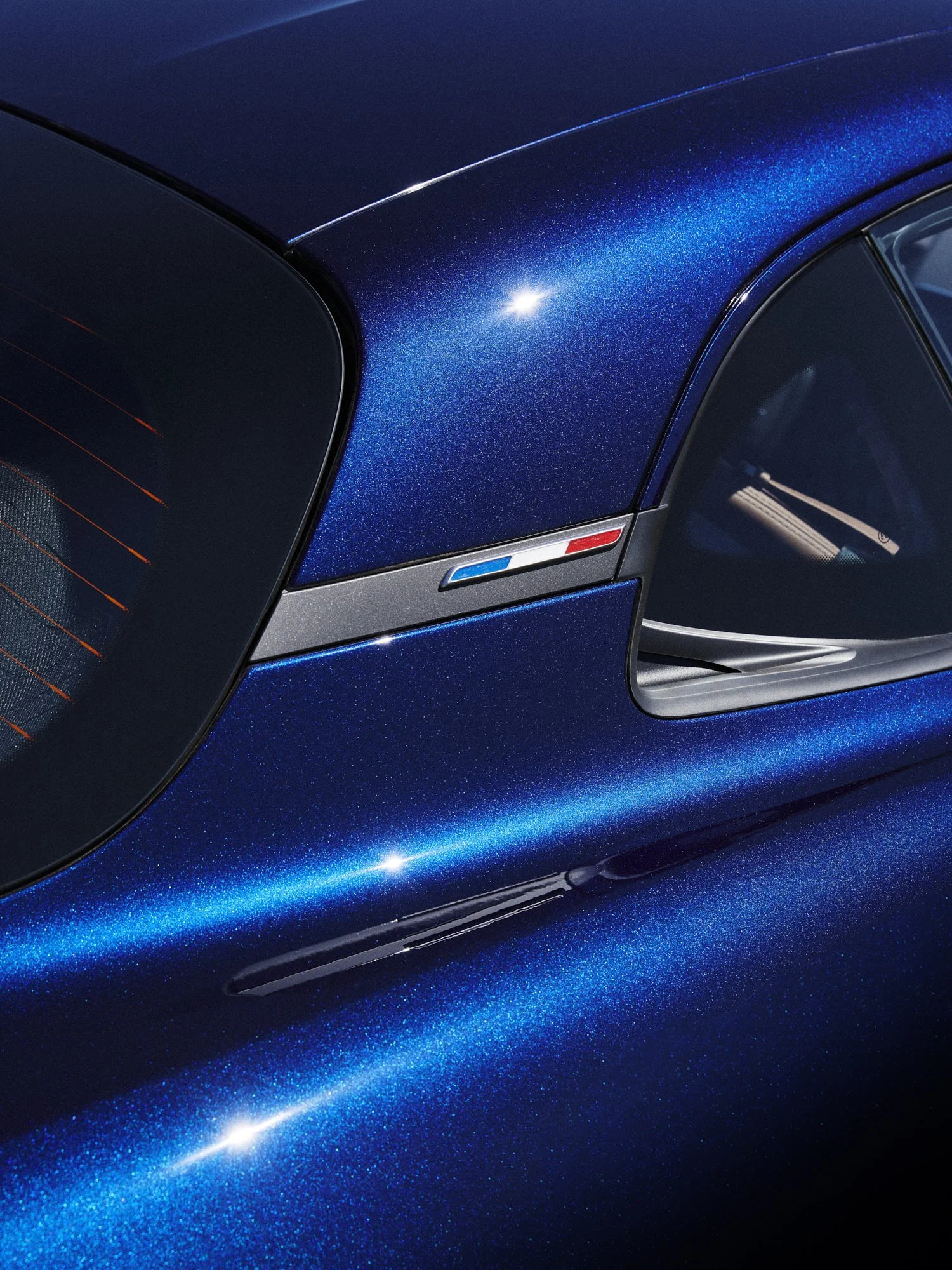Close-up of a blue sports car with a metallic finish, showing part of the rear window, side air intake, and a small strip of the rear light, emphasizing the glossy surface and sleek design.