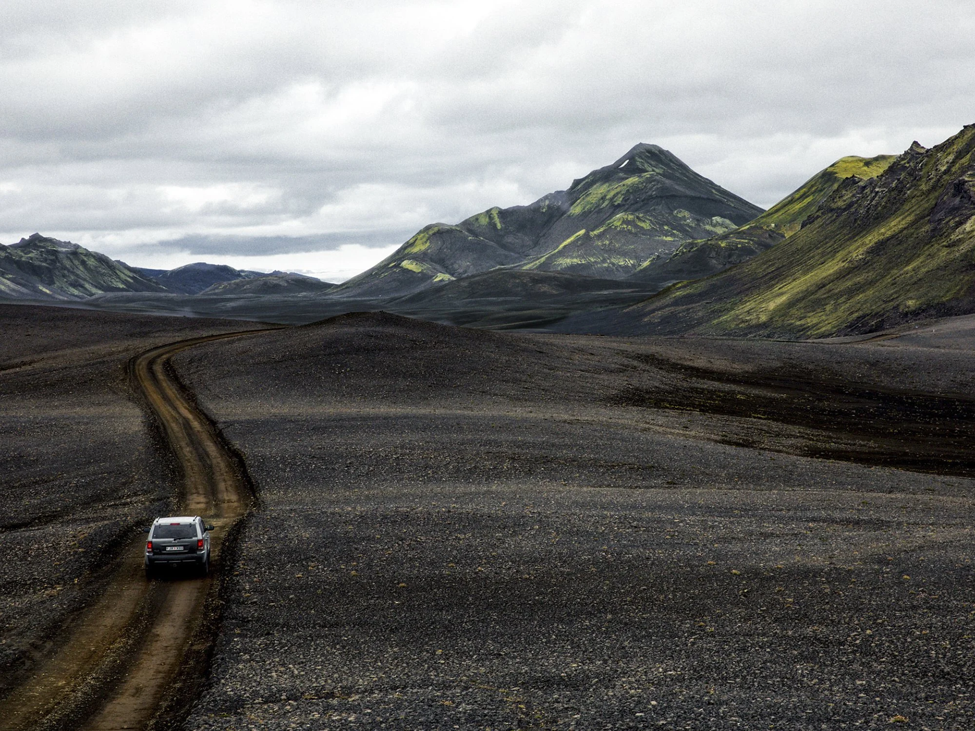 A car driving on a dirt road in a volcanic landscape with mountains covered in green moss under a cloudy sky.