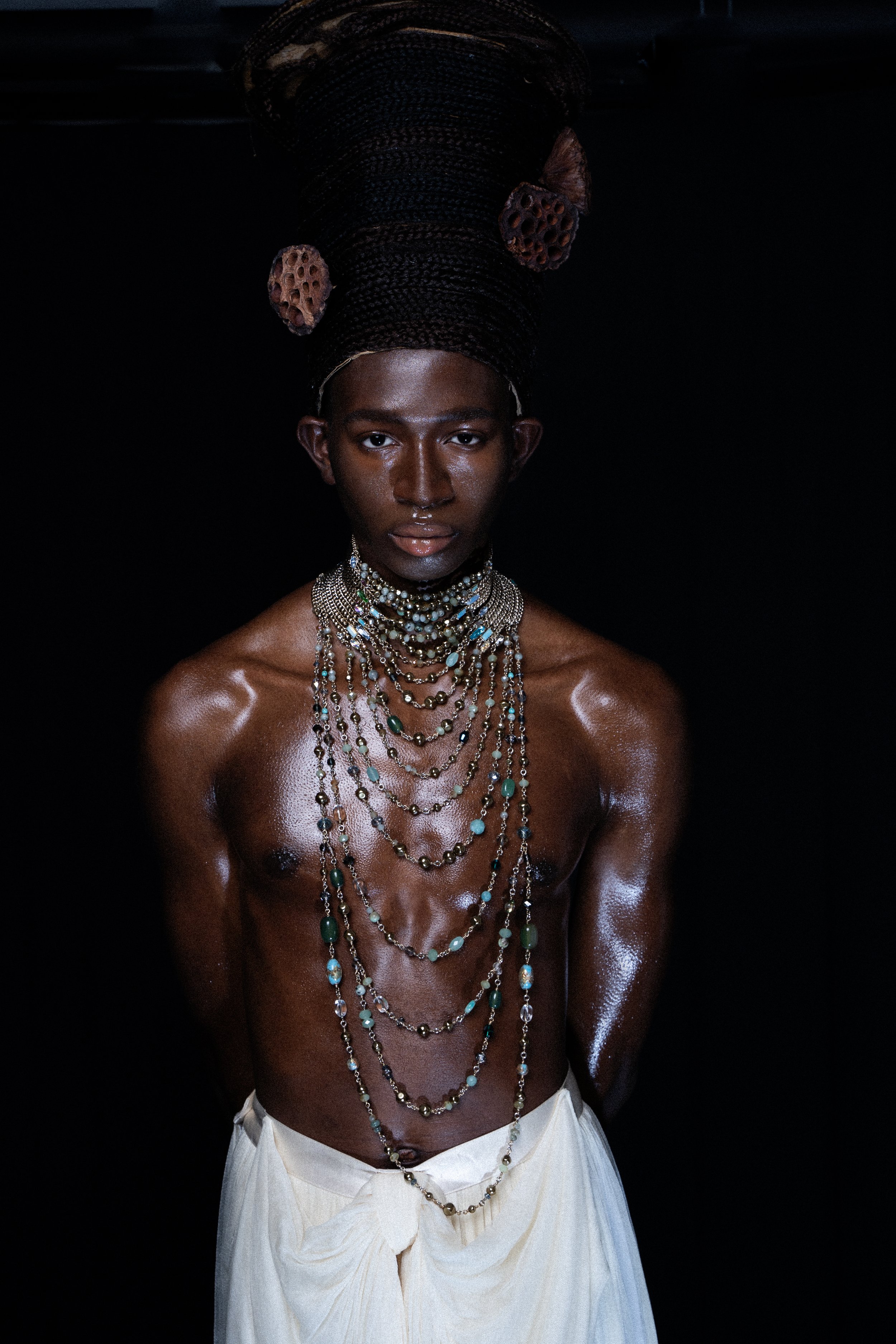 A person with dark skin and an elaborate headdress made of large coiled hair and lotus pods, standing against a dark background. They are shirtless, wearing multiple layered necklaces, and a white fabric draped around their waist.