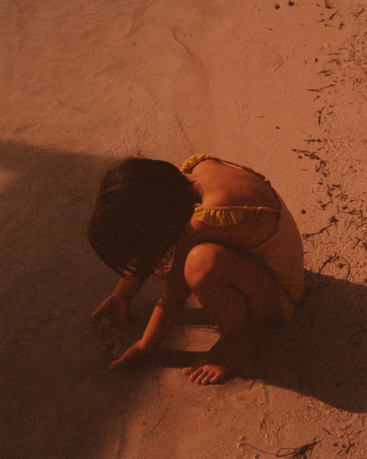 A child crouched on the ground at night, touching the dirt with her hand, with her head lowered and hair covering her face.