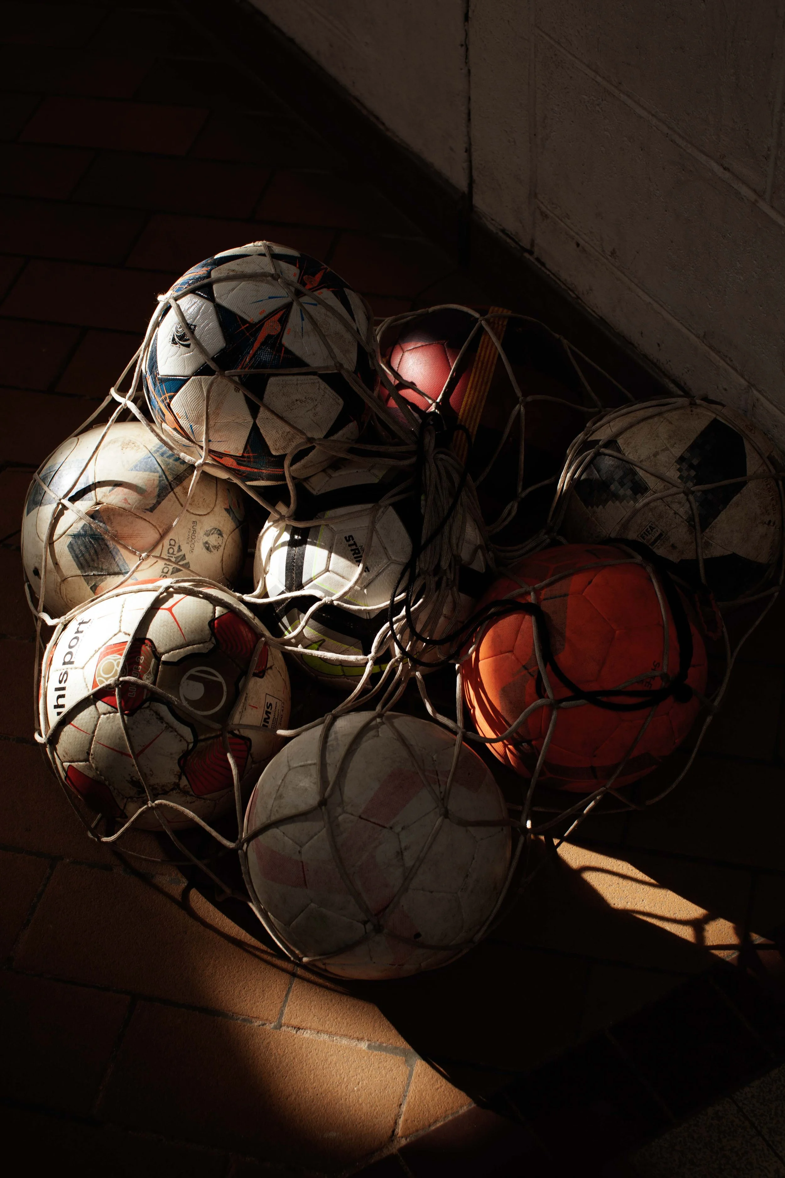 A tangled soccer ball net containing multiple used soccer balls, some with visible dirt, placed on a tiled floor with sunlight casting shadows.