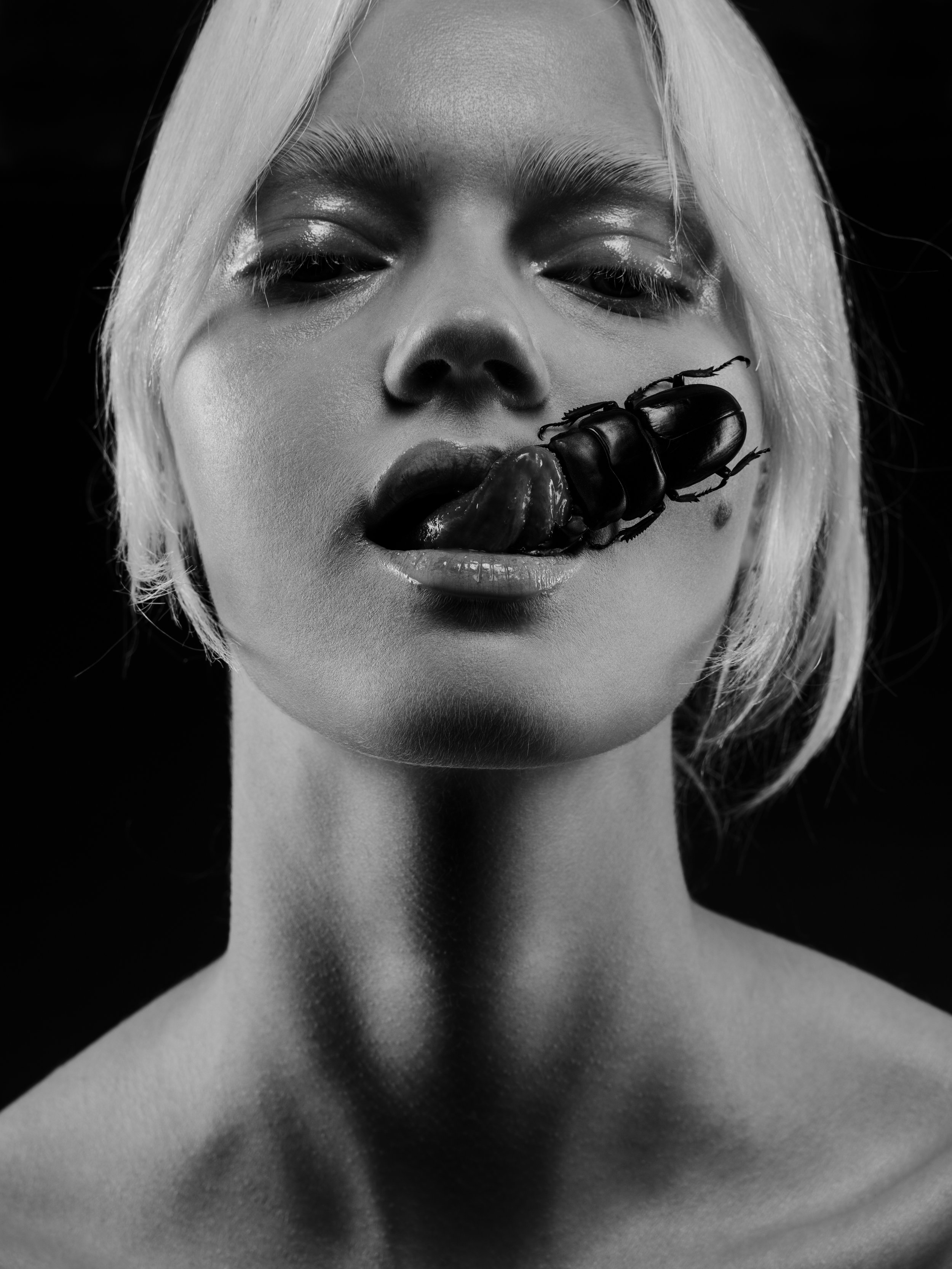 Black and white close-up portrait of a woman with short blonde hair, staring directly at the camera with her lips slightly parted. A large beetle is perched on her lips, with its mouthparts touching her tongue.