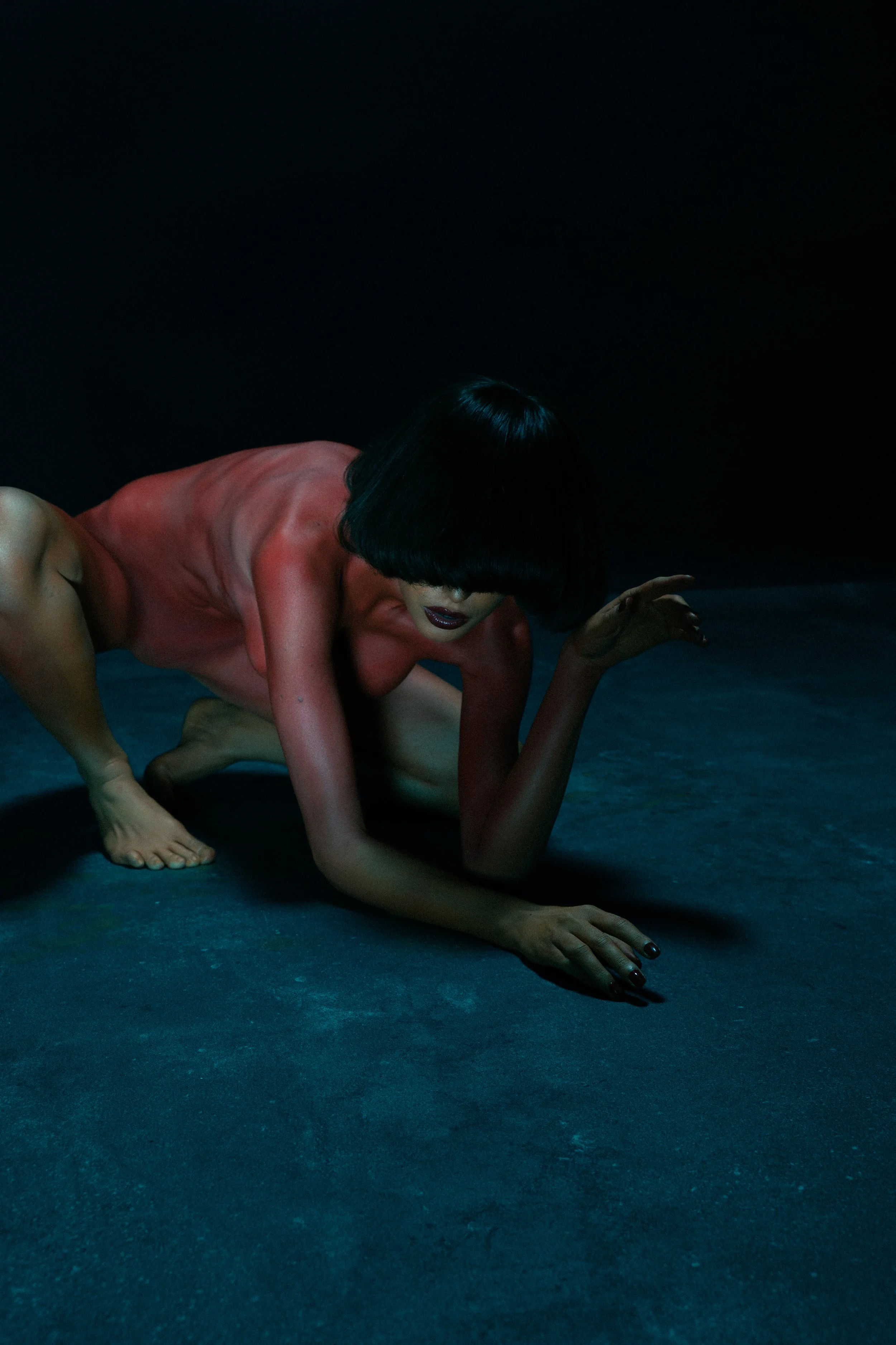 A person with black hair and dark lipstick crawling on a dark, textured floor in a dimly lit environment.