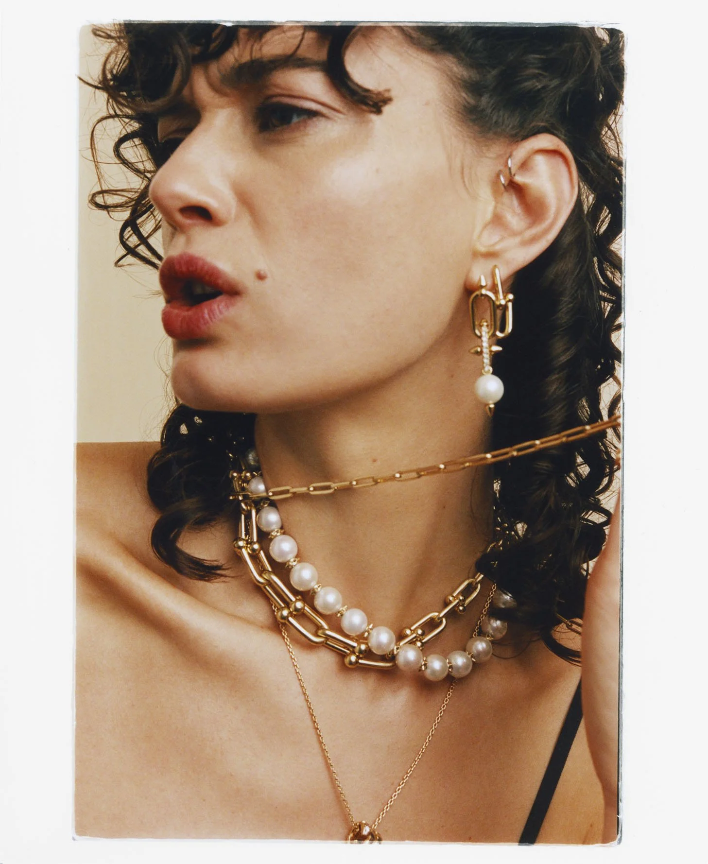 Close-up of a woman with curly dark hair wearing multiple gold and pearl jewelry necklaces and earrings, looking to the left.