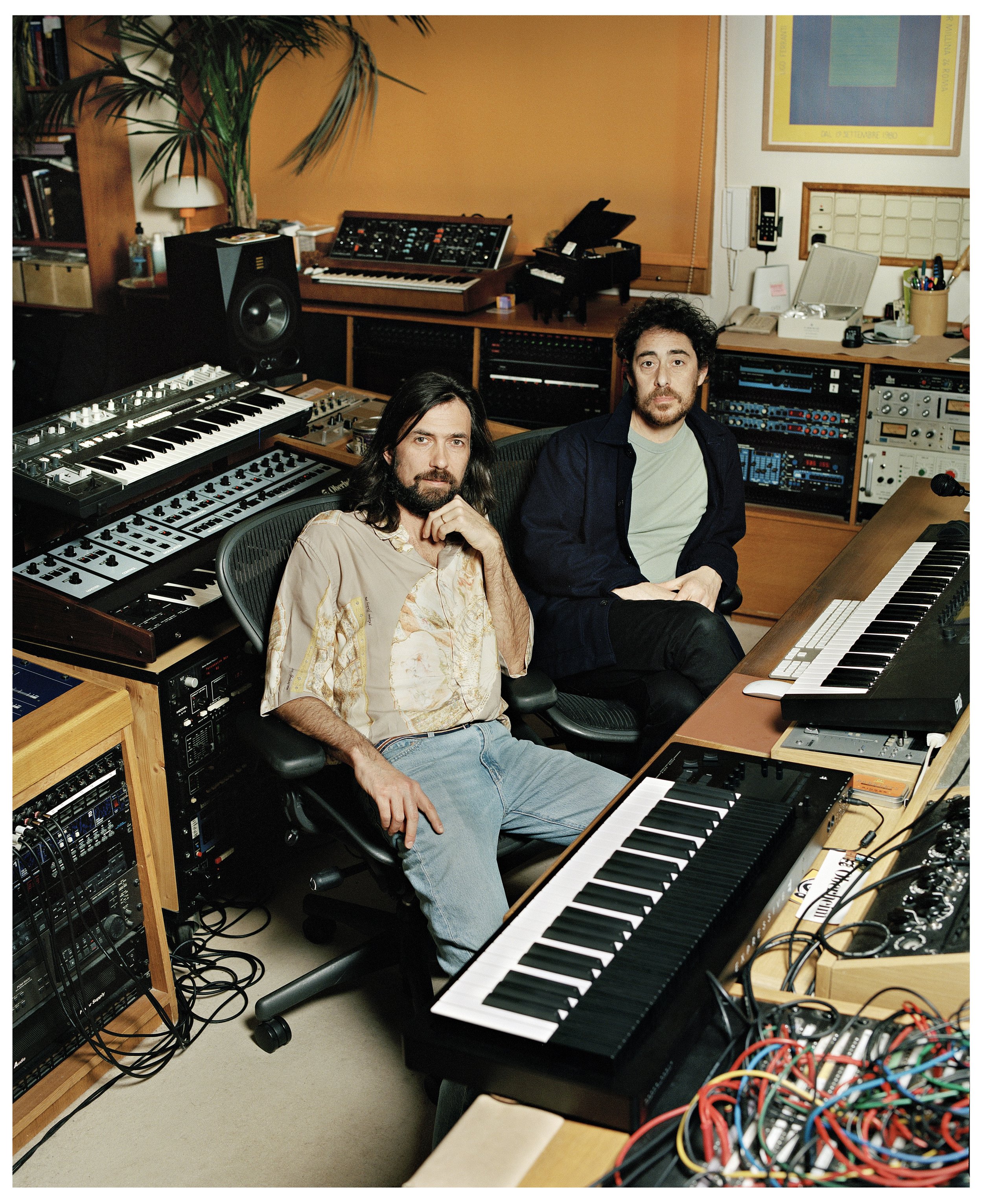 Two men in a recording studio surrounded by synthesizers, mixers, and other audio equipment.