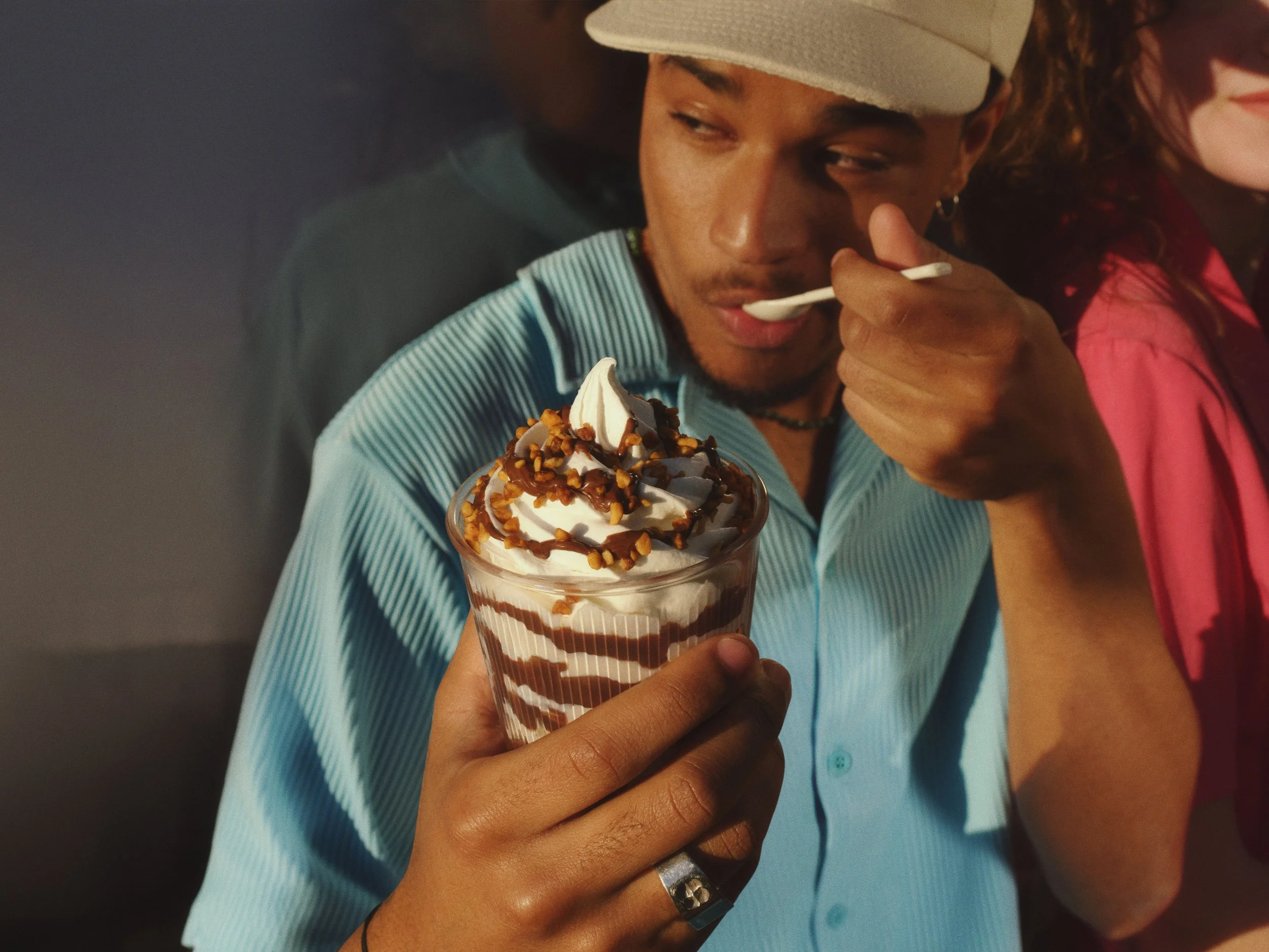 A man in a light blue shirt and white cap is holding a large glass of soft-serve ice cream topped with caramel and nuts, and is tasting it with a spoon.