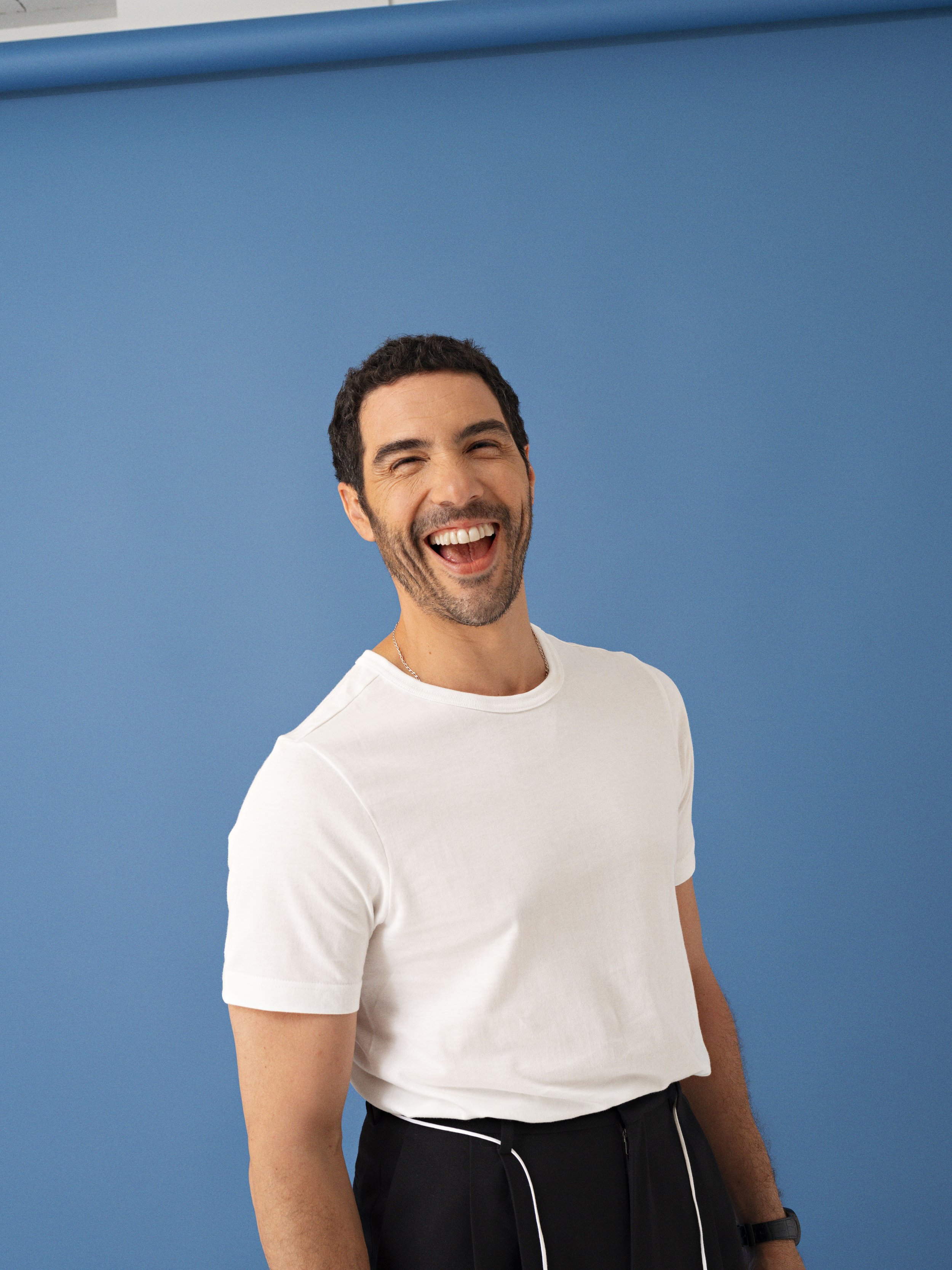 Man with dark hair and beard smiling and wearing a white t-shirt against a blue background.