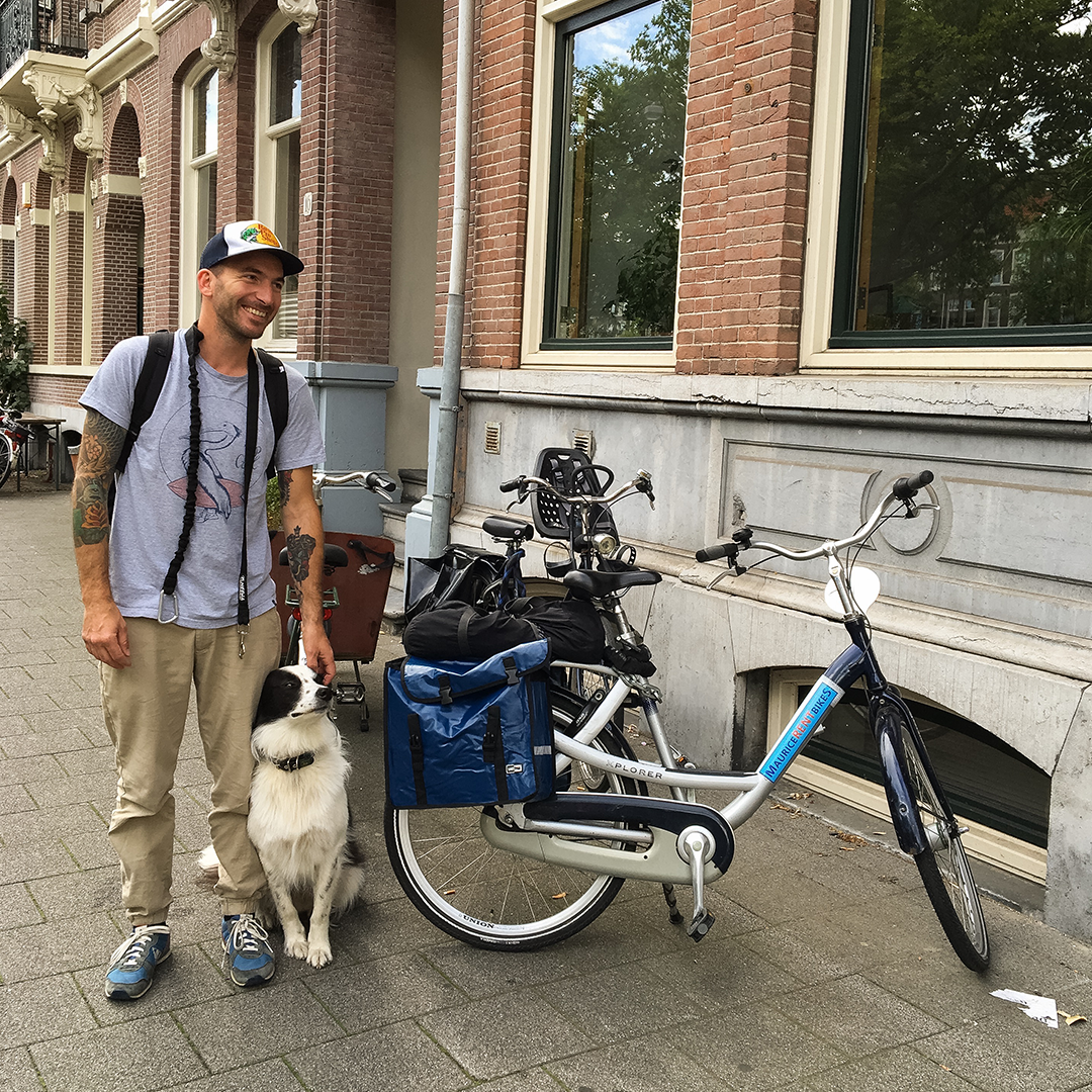 A man smiling with a black and white dog, standing next to a blue and white electric bike with luggage, on a city sidewalk in front of a brick building.