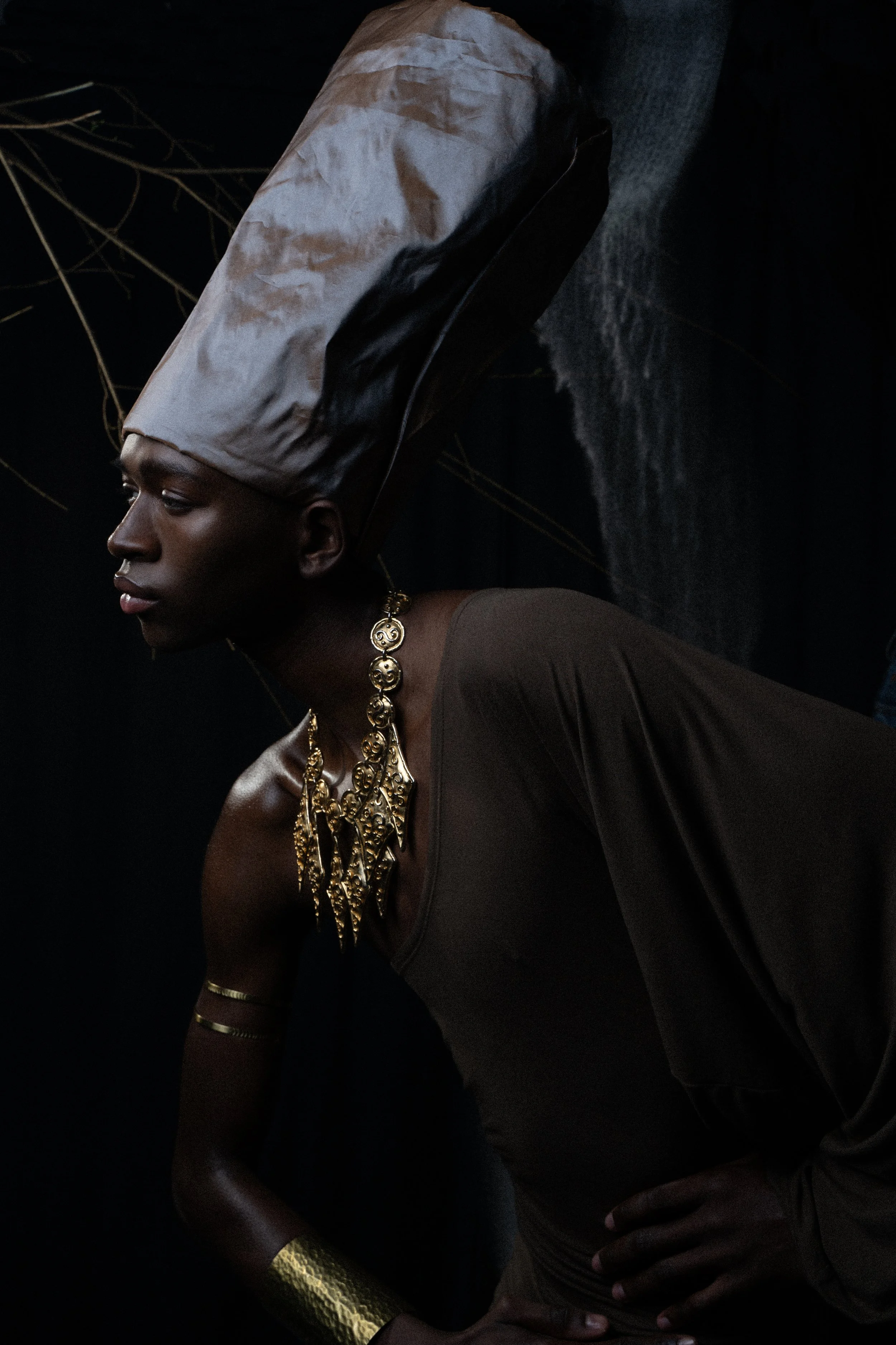 A woman dressed in dark clothing with gold jewelry and a large beige headpiece poses against a dark background.