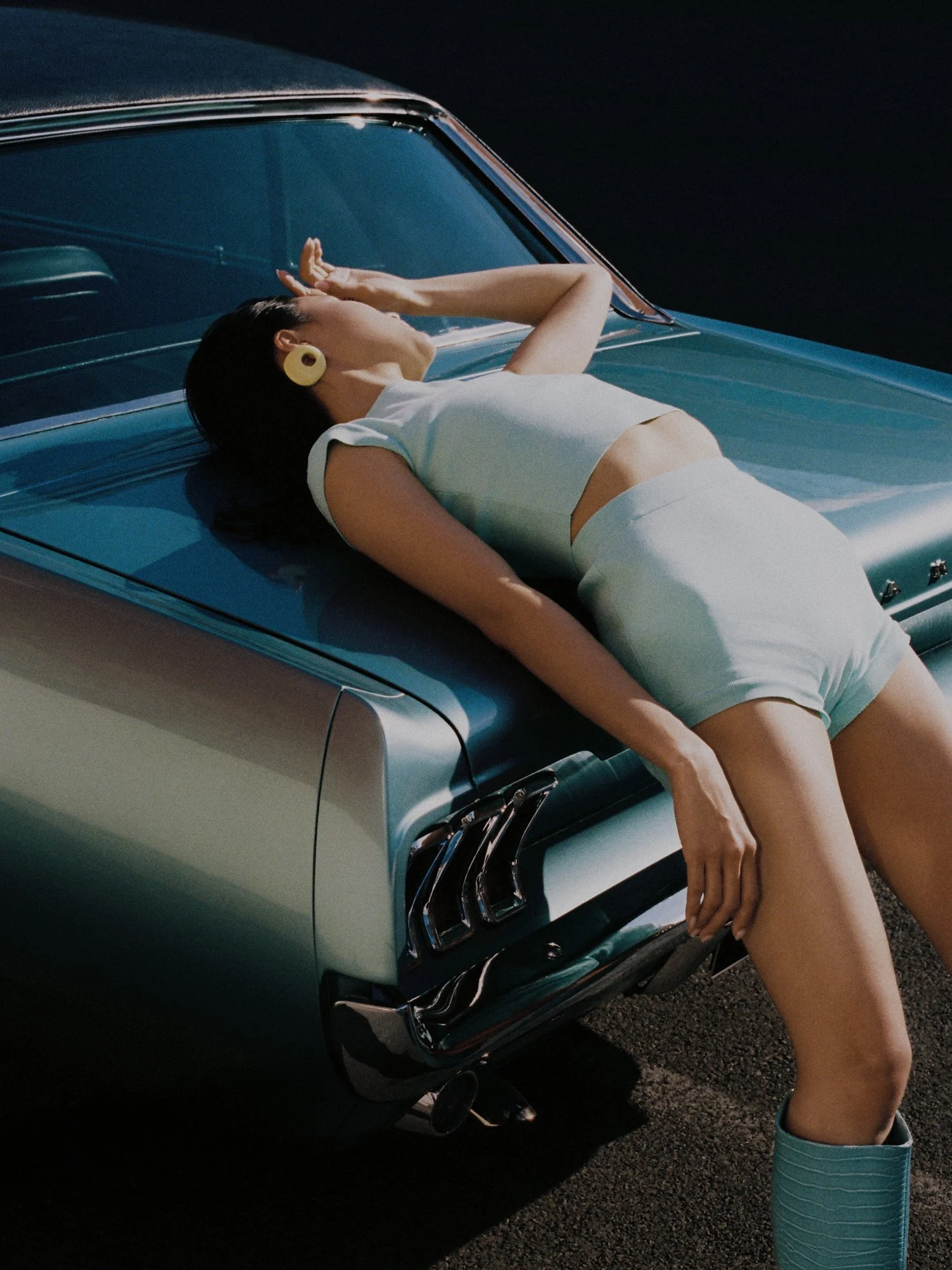 A woman lying on the hood of a vintage blue car, wearing a white outfit and large yellow earrings, with one arm resting on her forehead and the other hanging down, outdoors during daytime.