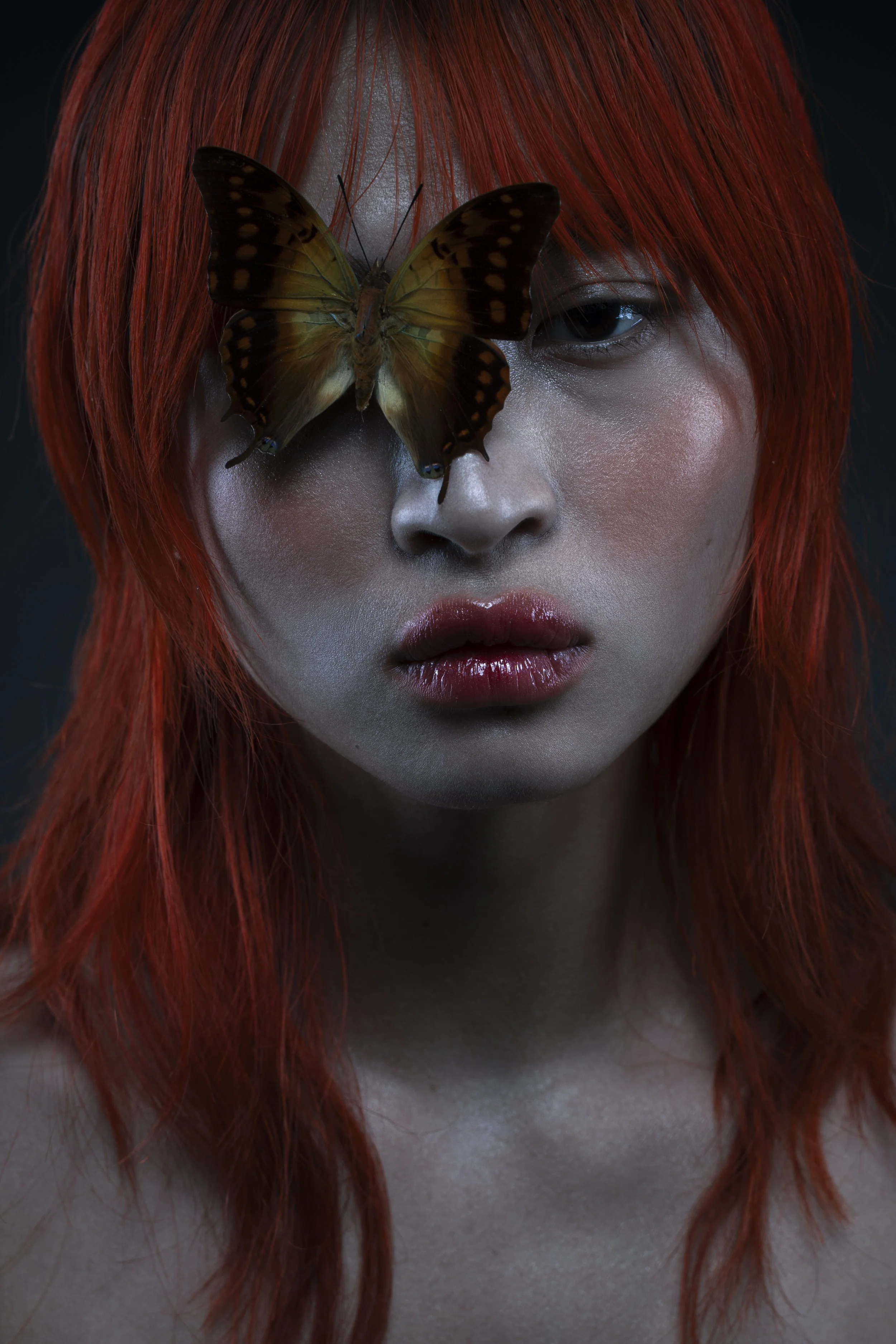 Close-up portrait of a woman with red hair, makeup, and a butterfly resting on her face near her eye.