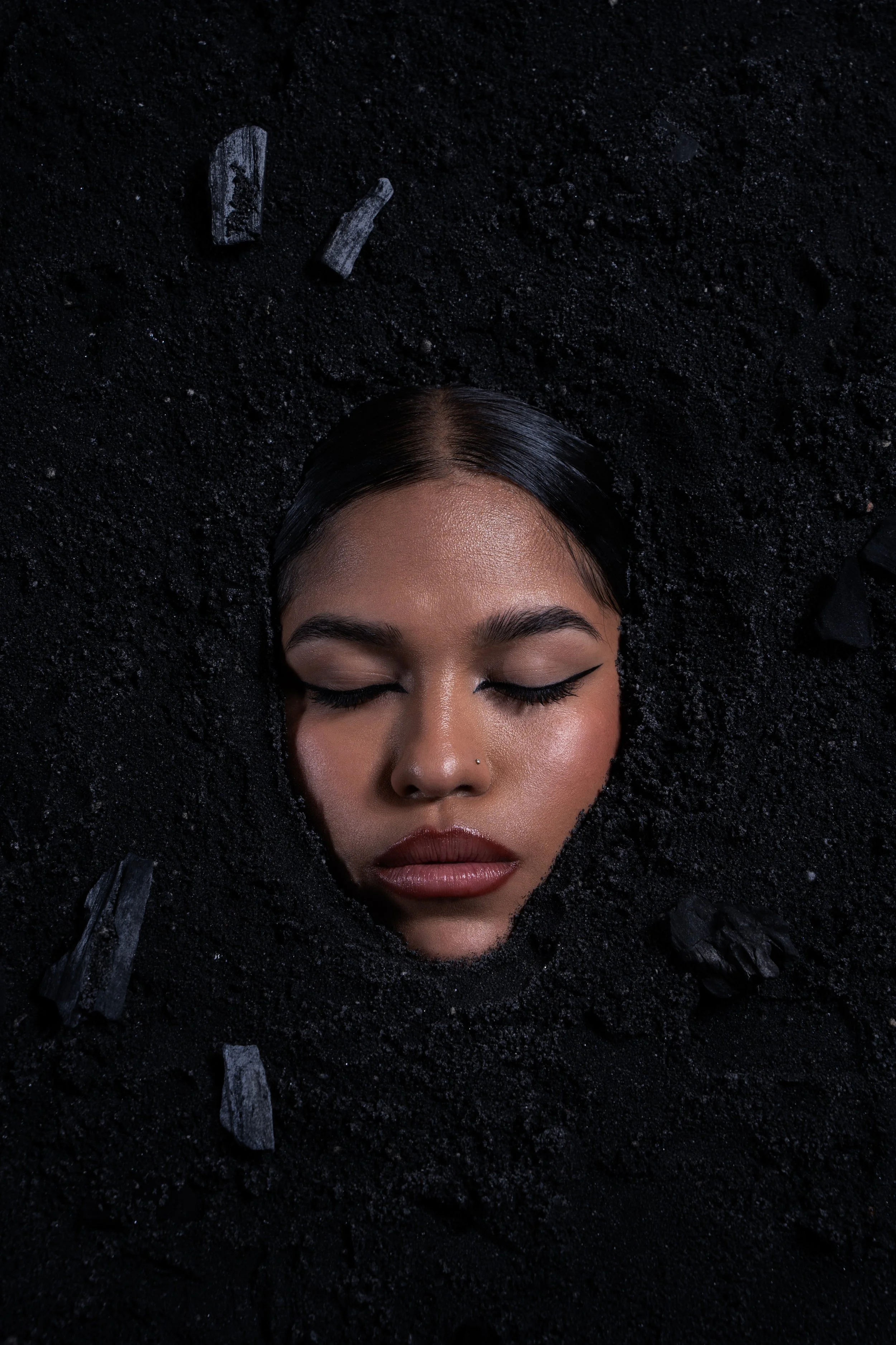 A woman with closed eyes and dark makeup is partially submerged in black sand.