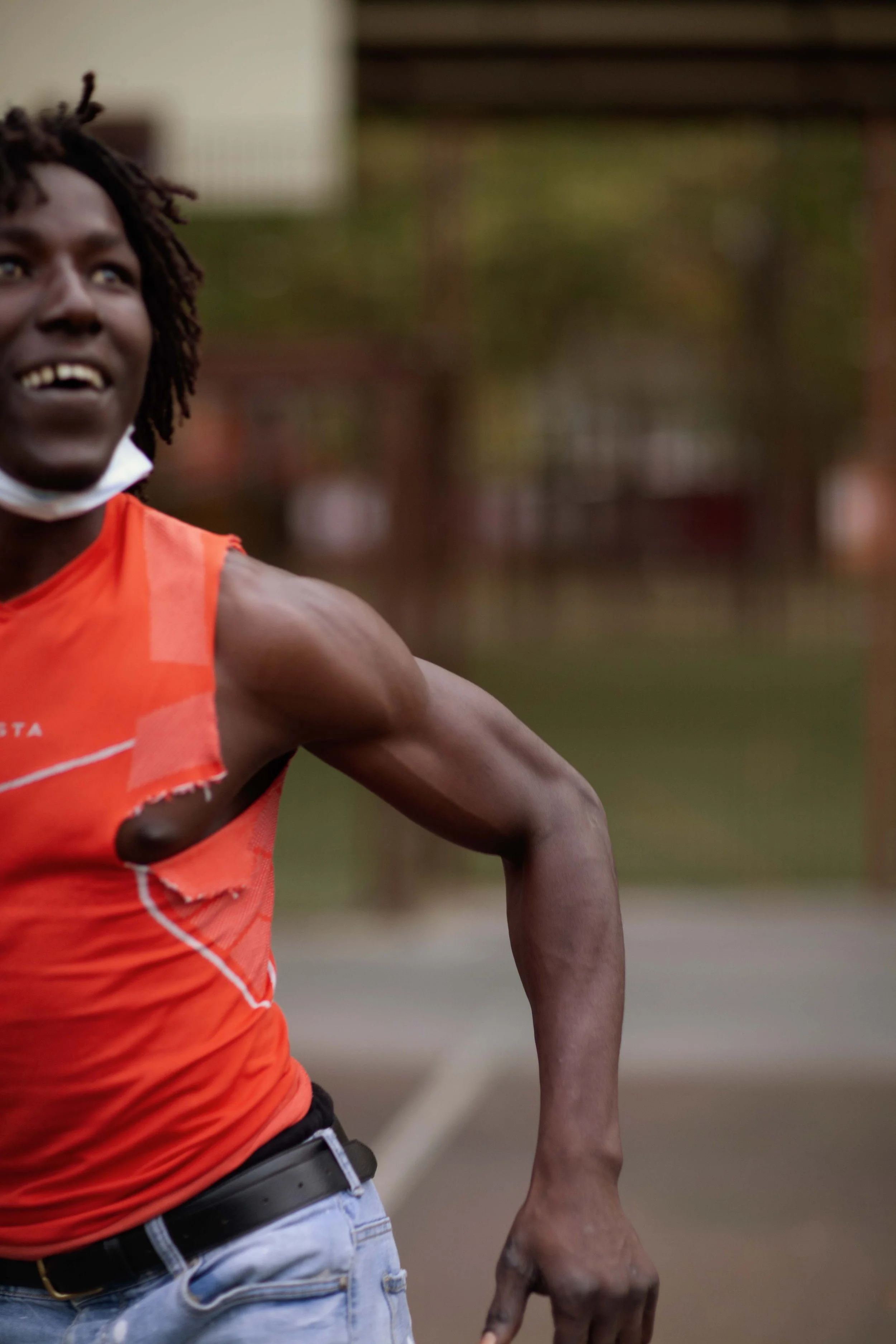 A person running outdoors, wearing a torn orange tank top and jeans, with a mask pulled down under their chin, smiling, on a sports court with a blurred background.