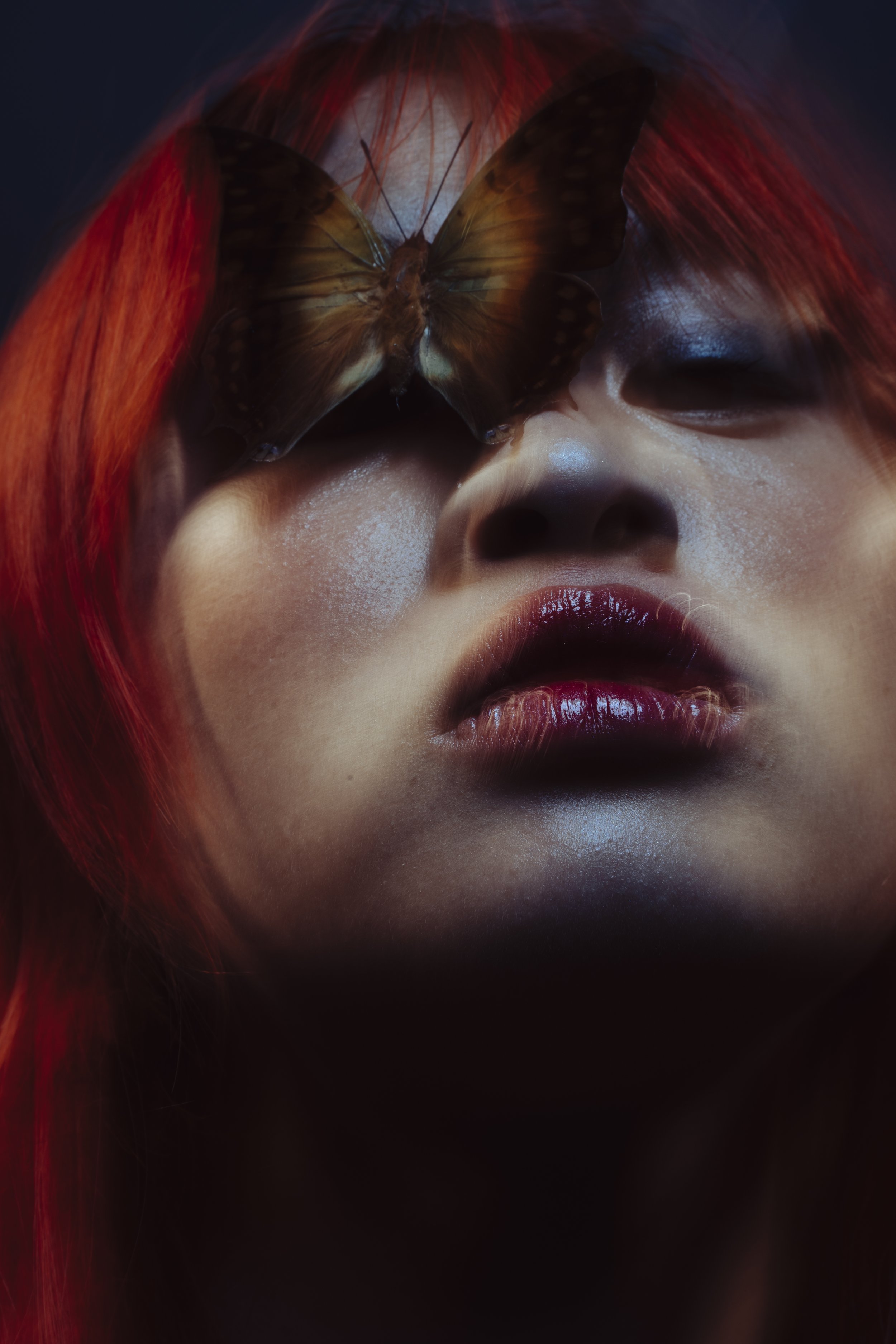 Close-up of a woman with red hair, closed eyes, and glossy lips, with a butterfly resting on her forehead.
