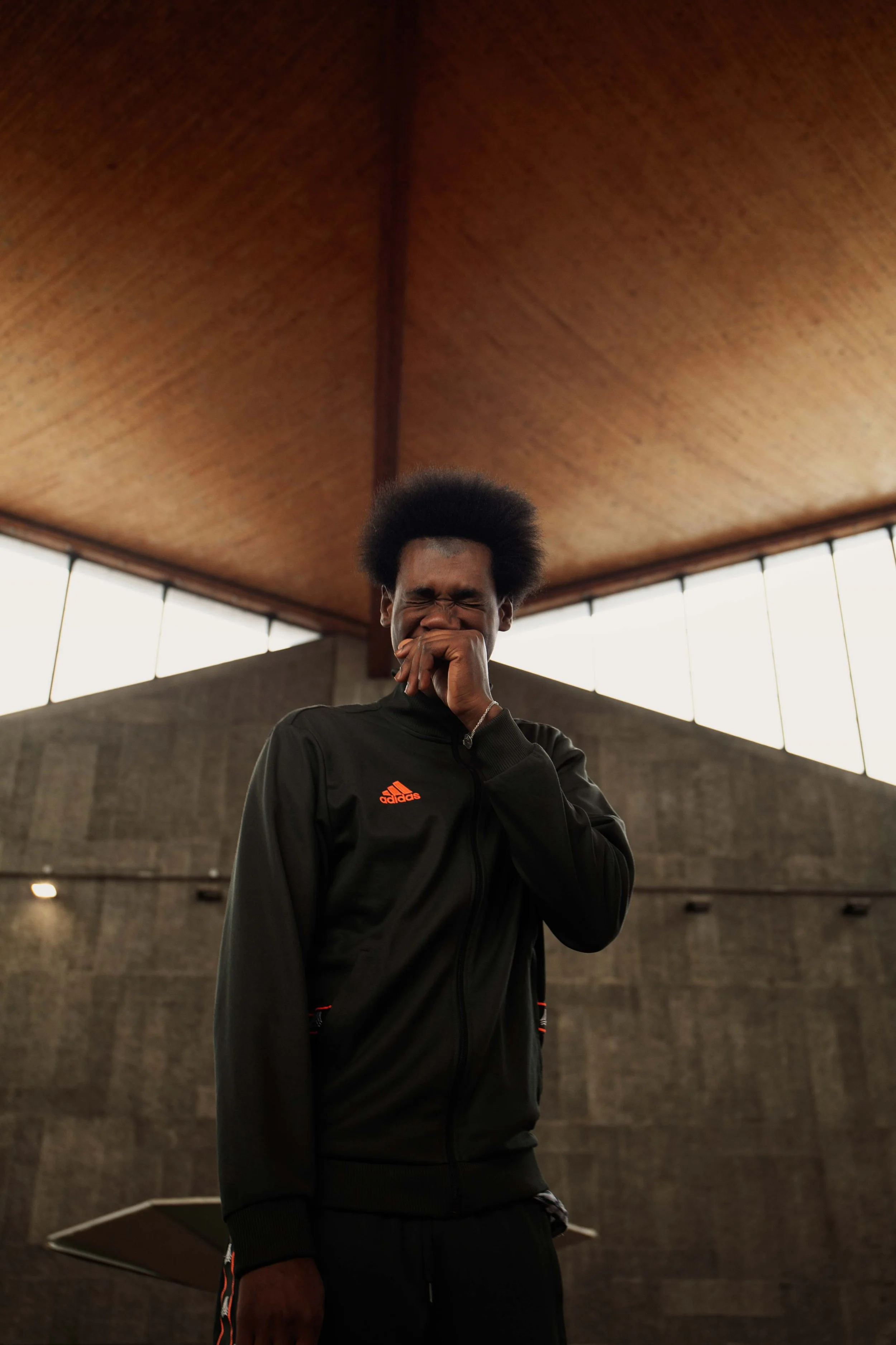 A young man with a natural afro hairstyle in a dark sports jacket, standing indoors with his hand covering his mouth while smiling or laughing.