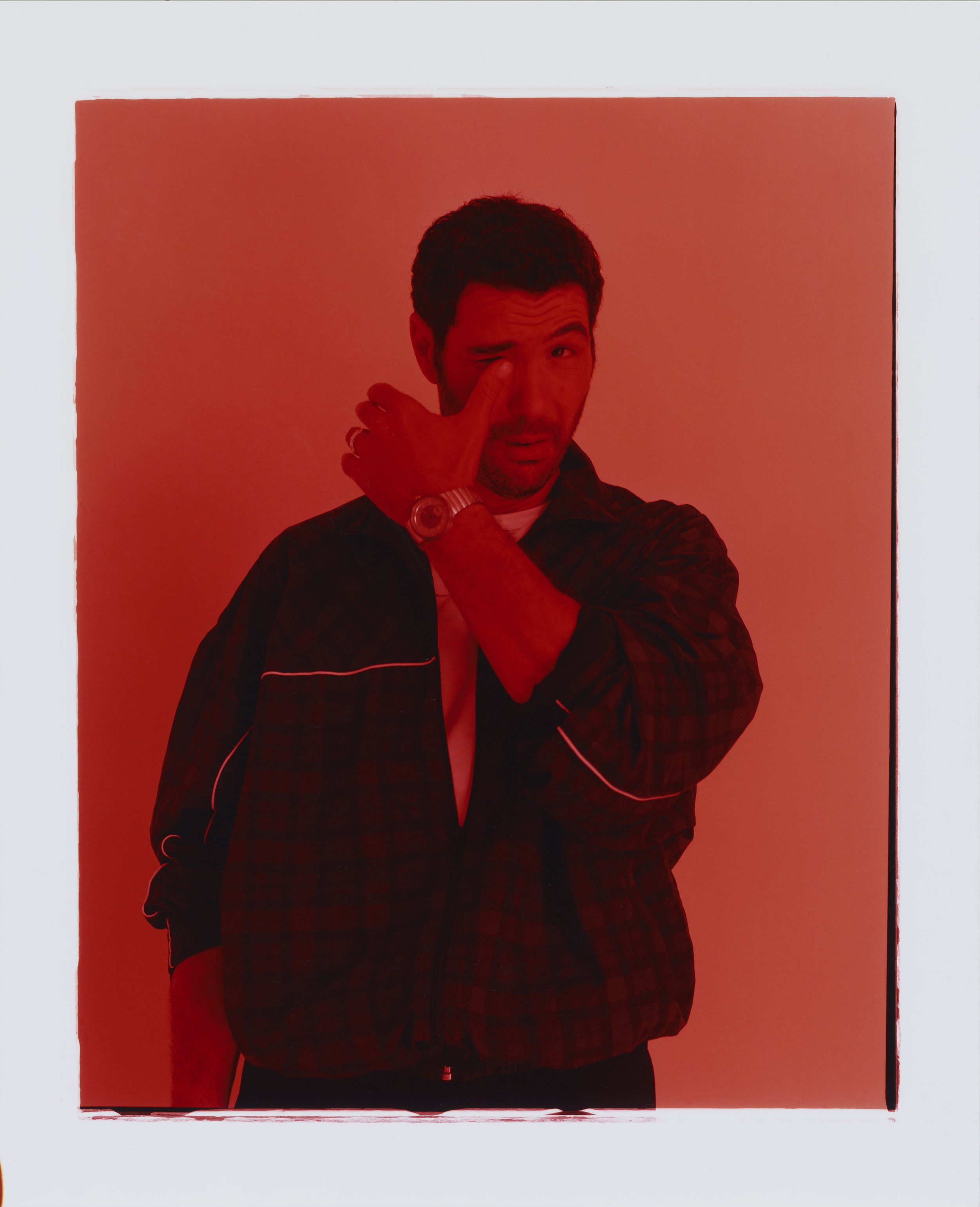 Man with short dark hair in a black plaid jacket making a gesture with one eye closed and one eye open, standing against a red background.