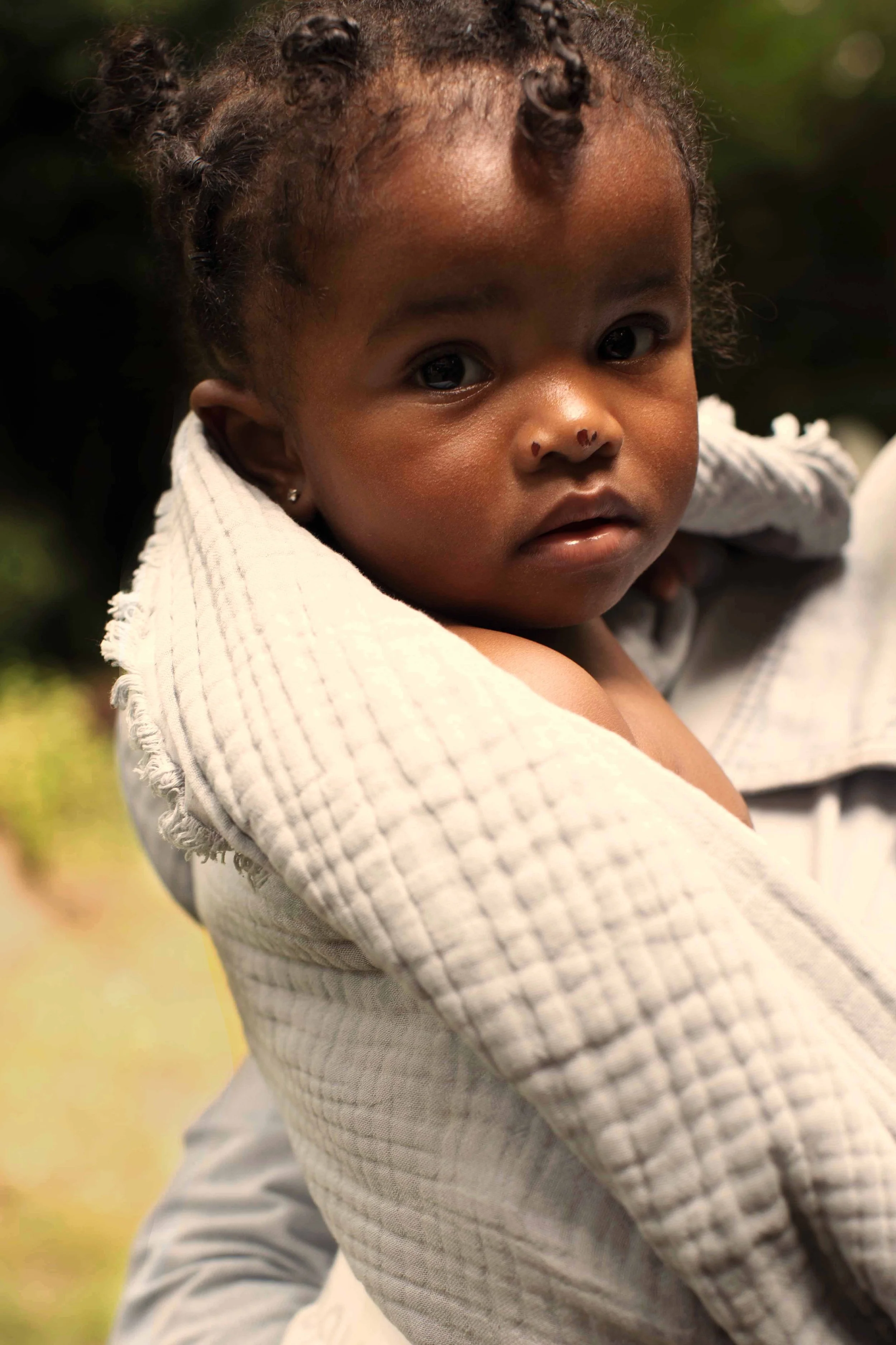 A young child with dark skin and curly hair styled in small twists, wrapped in a light-colored quilted blanket, looking directly at the camera with a curious expression.