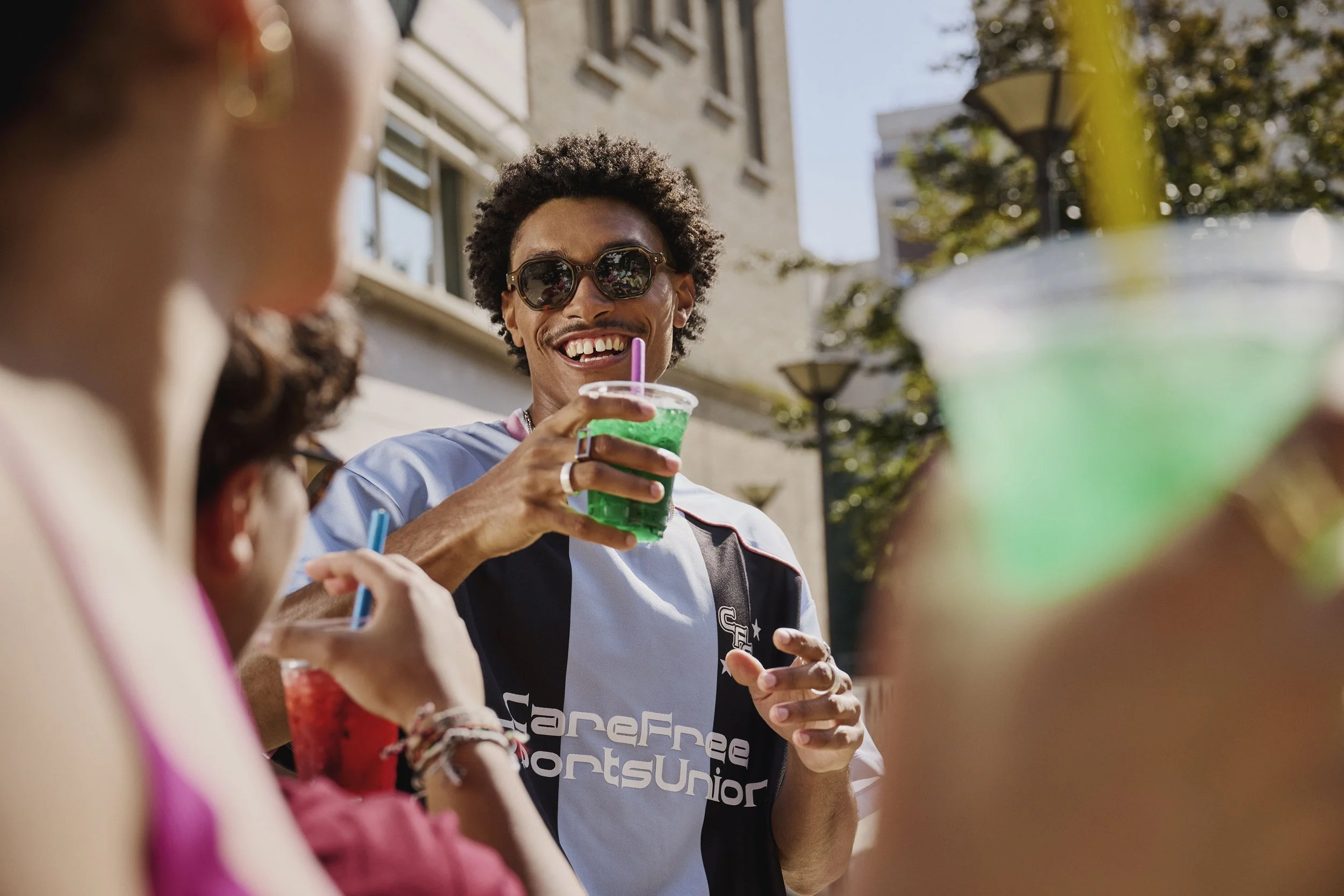 A man with curly hair, sunglasses, and a broad smile holding a green drink with a straw, engaging in conversation with others outdoors on a sunny day near buildings and trees.