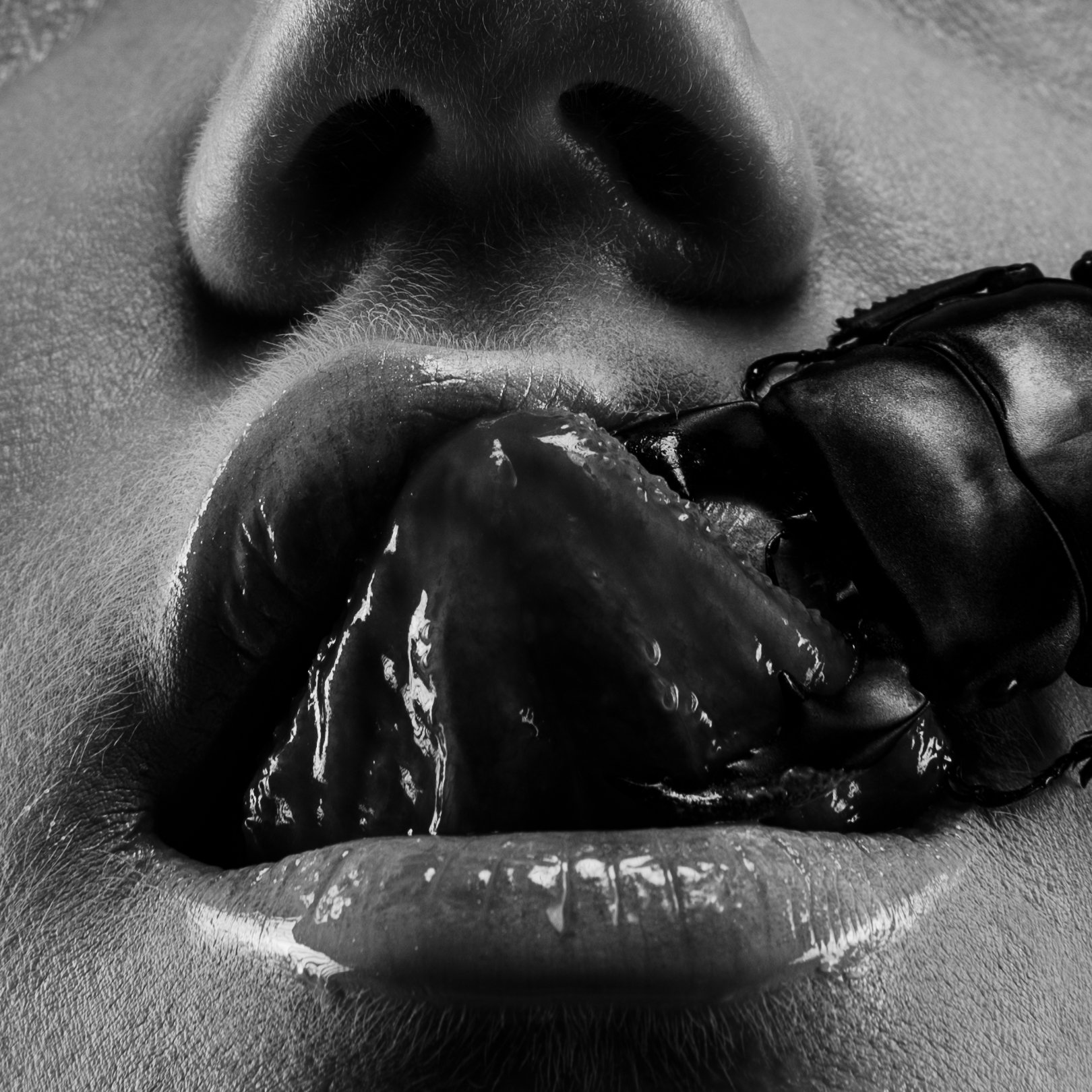 Close-up black and white photo of a person's face, with their lips parted and tongue sticking out, touching a gloved hand holding a smooth stone.