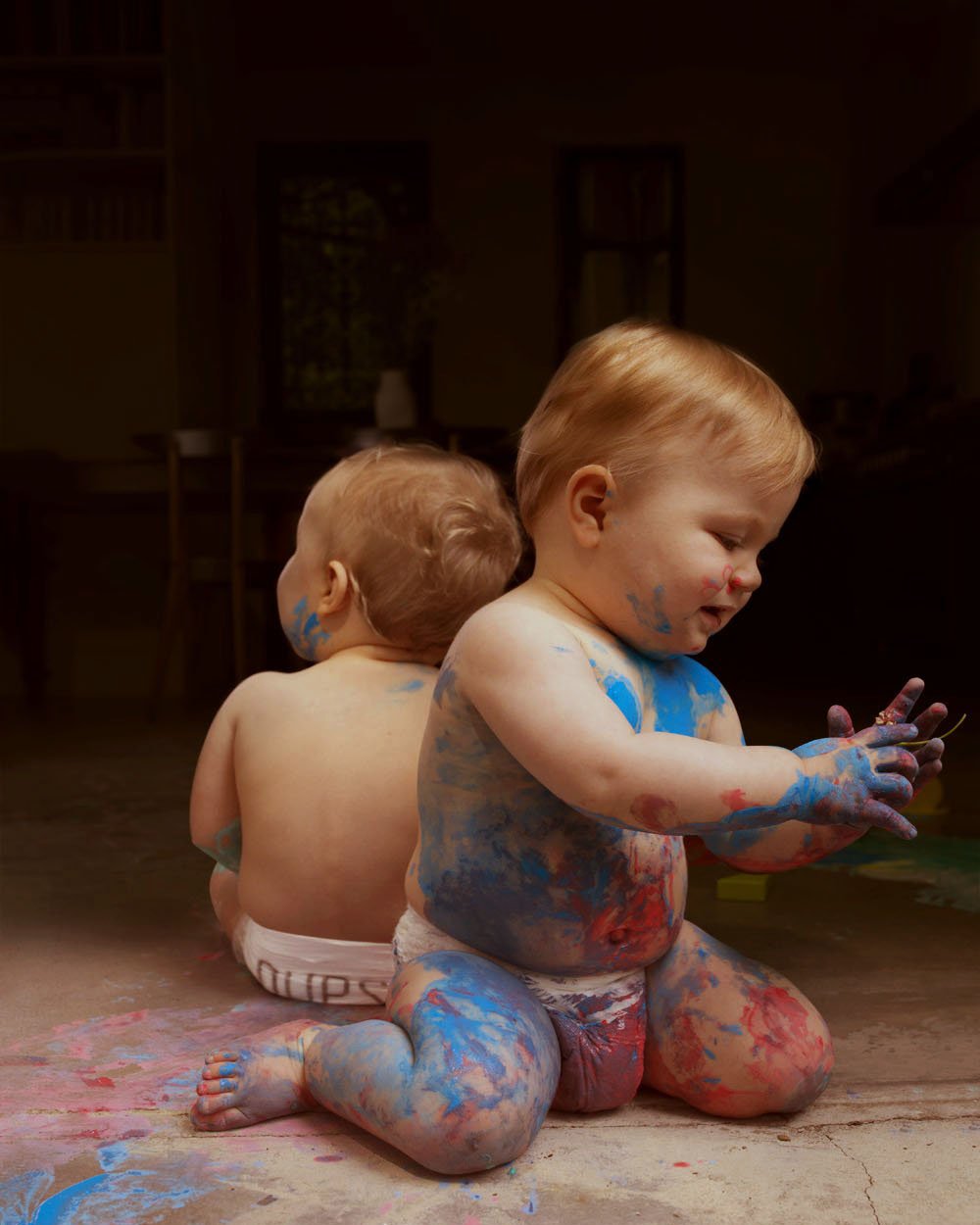 Two young children with paint-covered bodies sitting on the floor of a dimly lit room, playing and engaging with their hands.