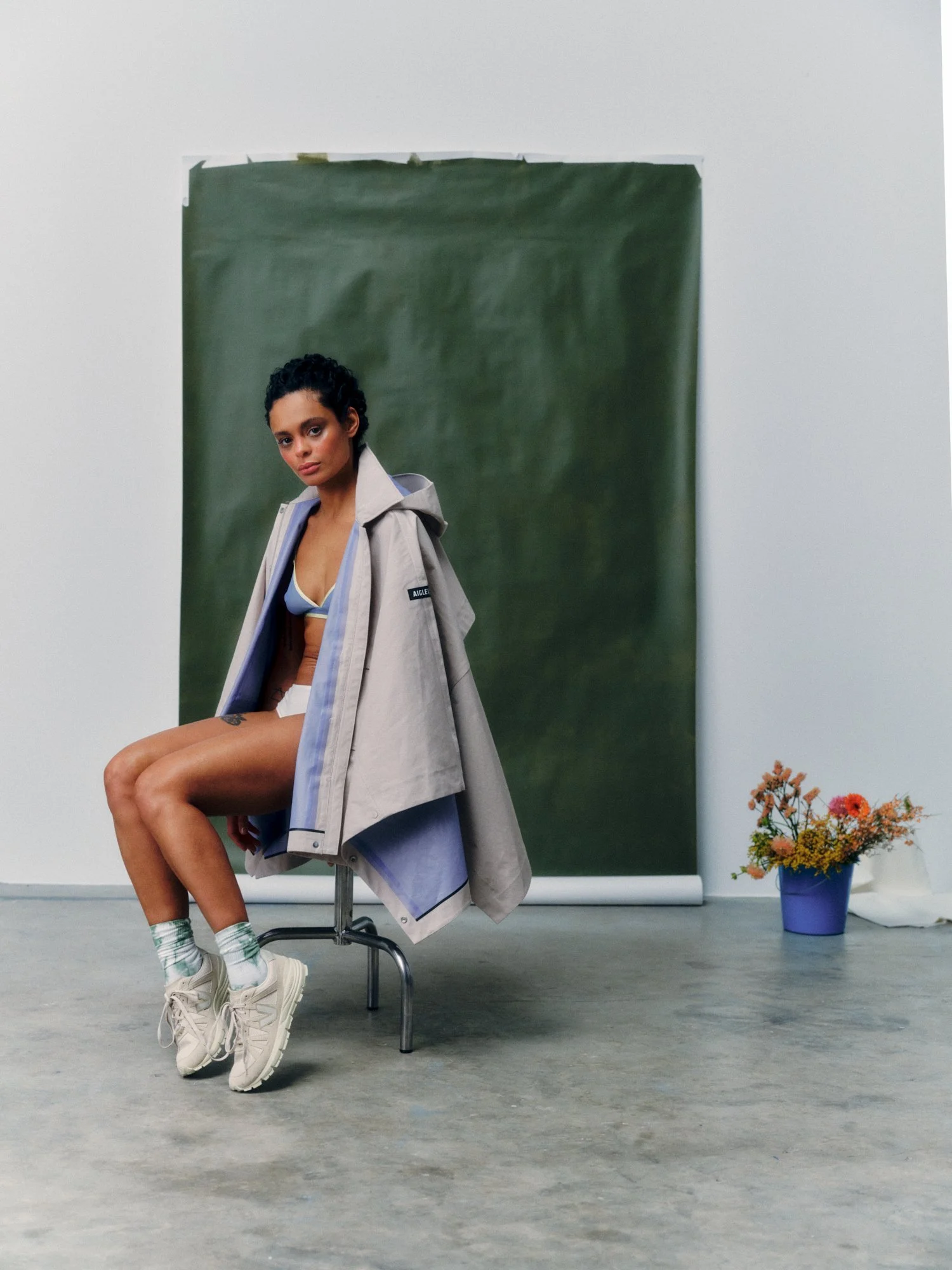 A young woman with short curly black hair and tan skin sitting on a chair in front of a green backdrop. She is wearing a beige jacket over blue sportswear and white sneakers with striped socks. A blue flowerpot with orange and yellow flowers is on th
