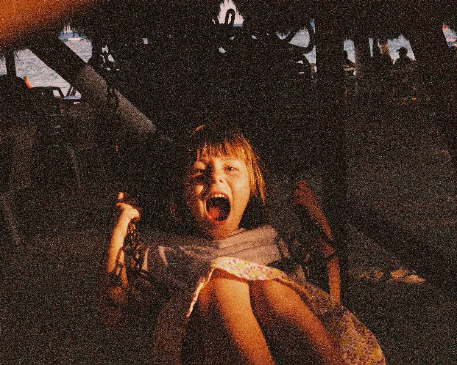 A young girl with short brown hair and bangs, sitting on a swing, yelling with her mouth wide open, during sunset at a beach or waterfront location.