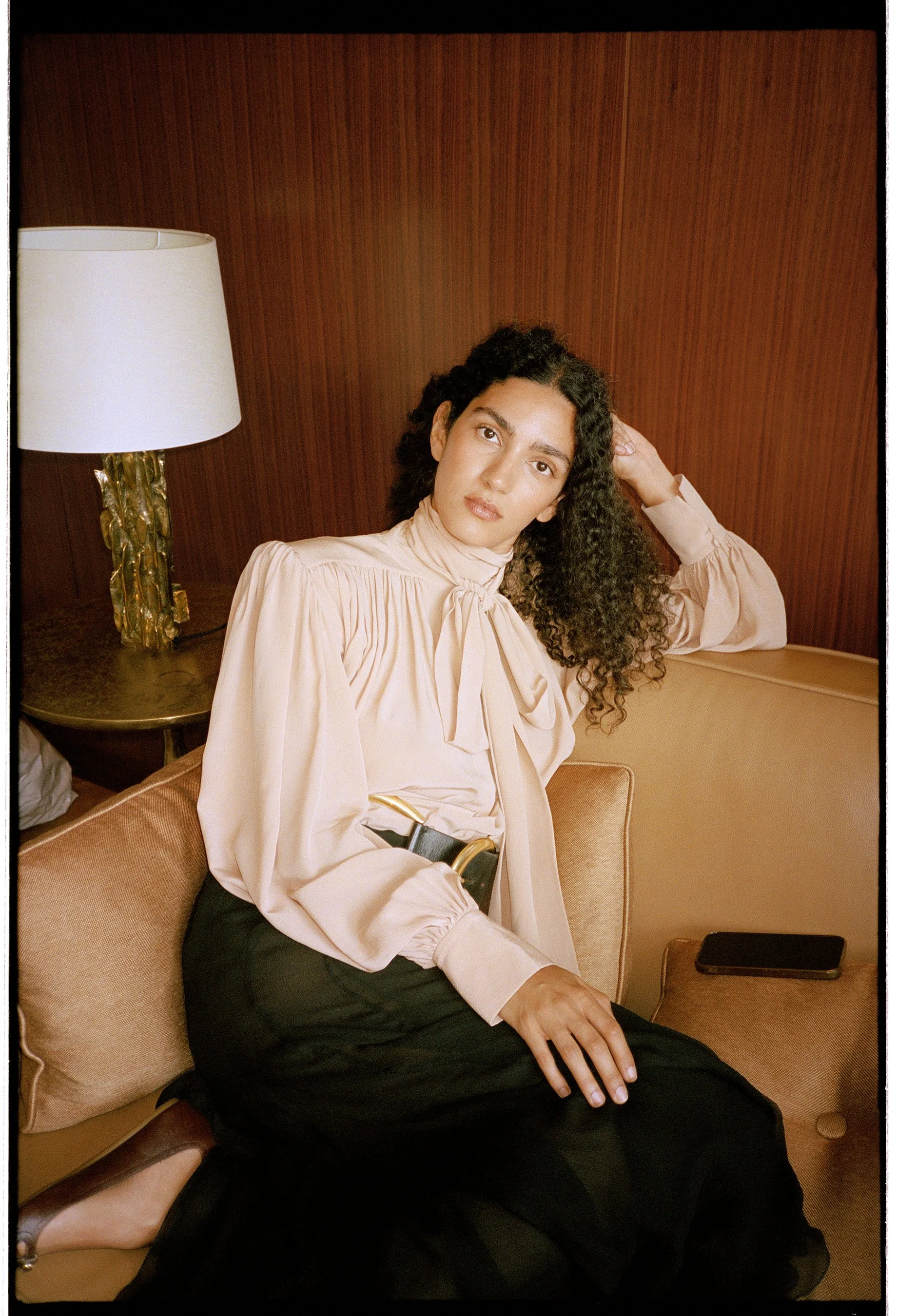 A young woman with curly dark hair sitting on a beige sofa in a room with wooden walls, wearing a light pink blouse with a bow tie at the neck, and black pants. She has a serious expression and is looking at the camera, with her left hand resting on 
