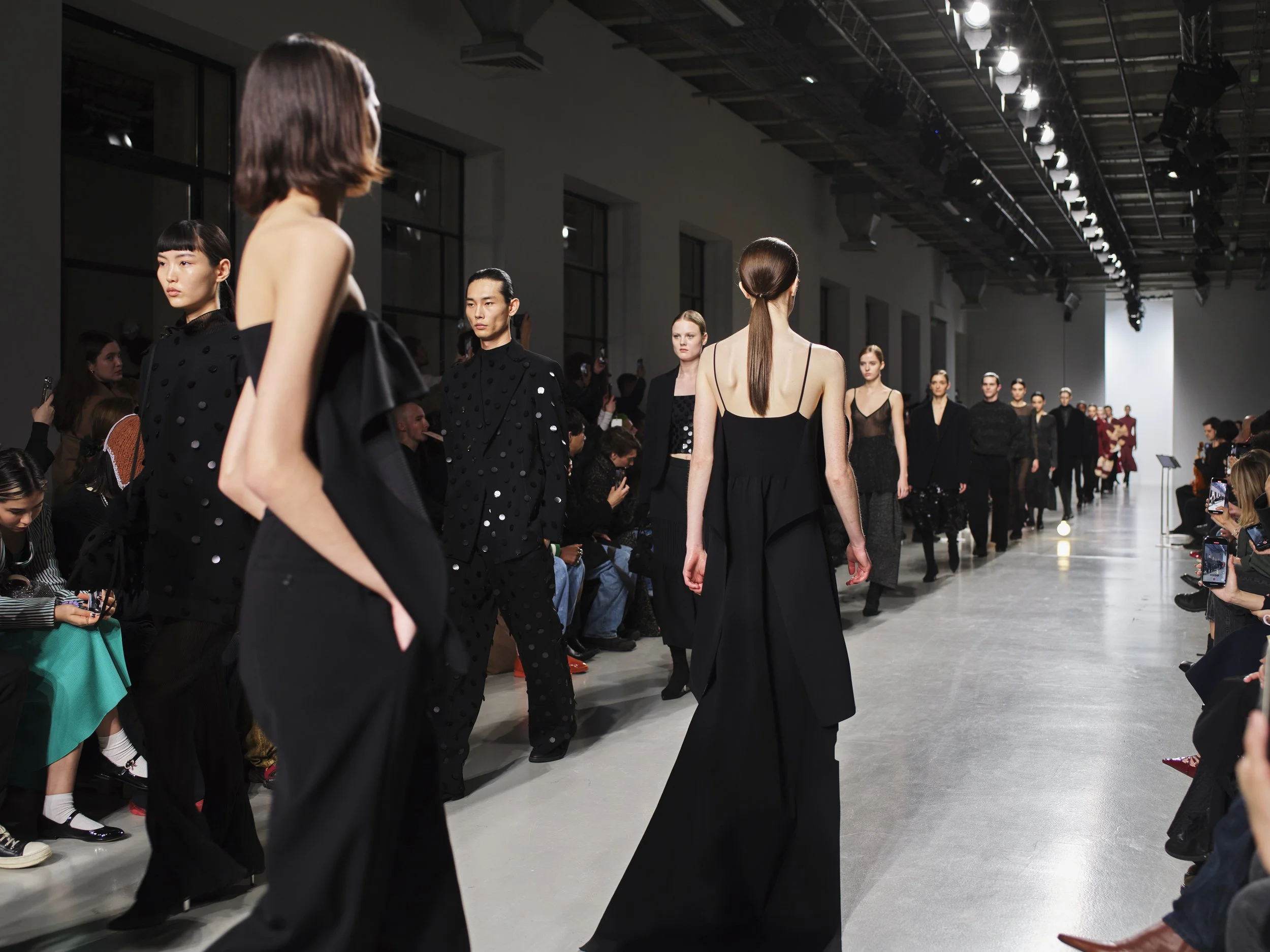 Models walking down the runway during a fashion show, with audience members seated on either side taking photos.