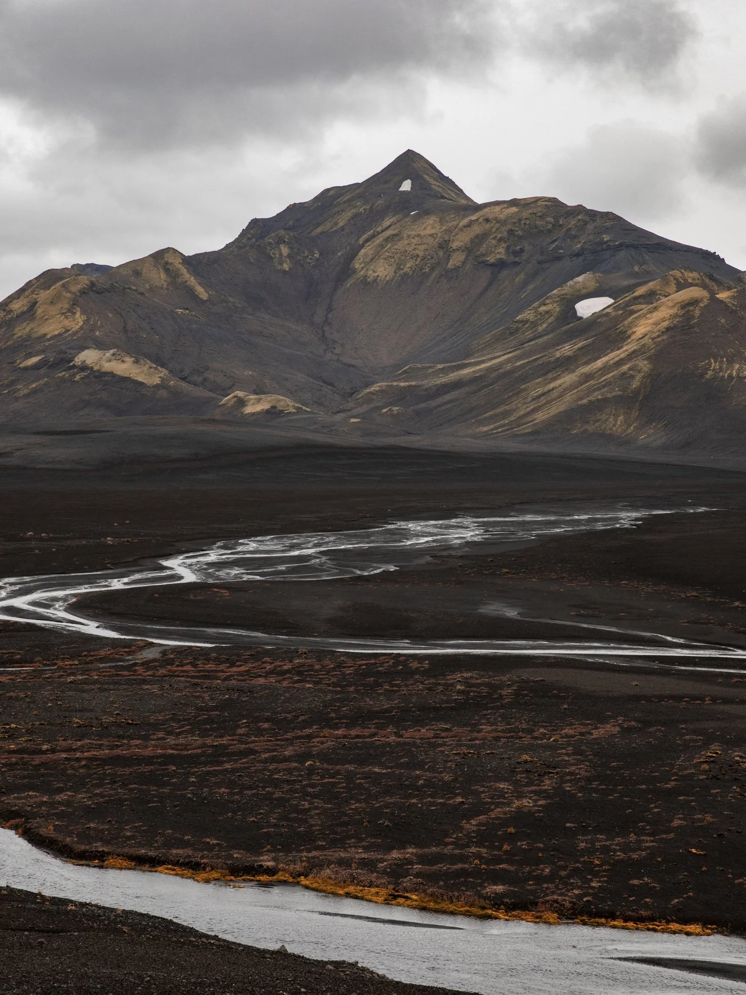 A dark, volcanic mountain with patches of snow on its slopes under a cloudy sky, with a winding river or stream in the foreground.